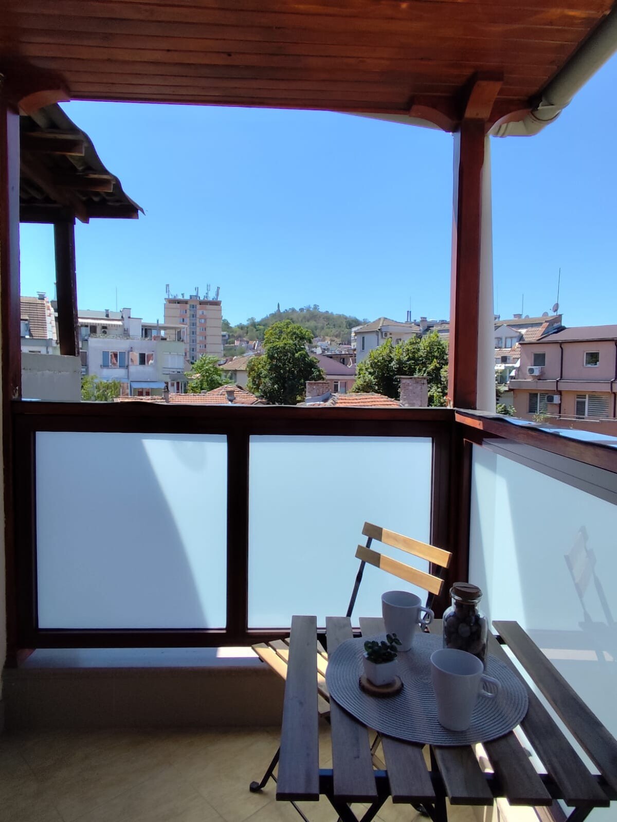 A small terrace offers a wooden table with two chairs. A scenic view of Alyosha hill is visible beyond the balcony's glass railing, framed by clear blue skies and surrounding rooftops.