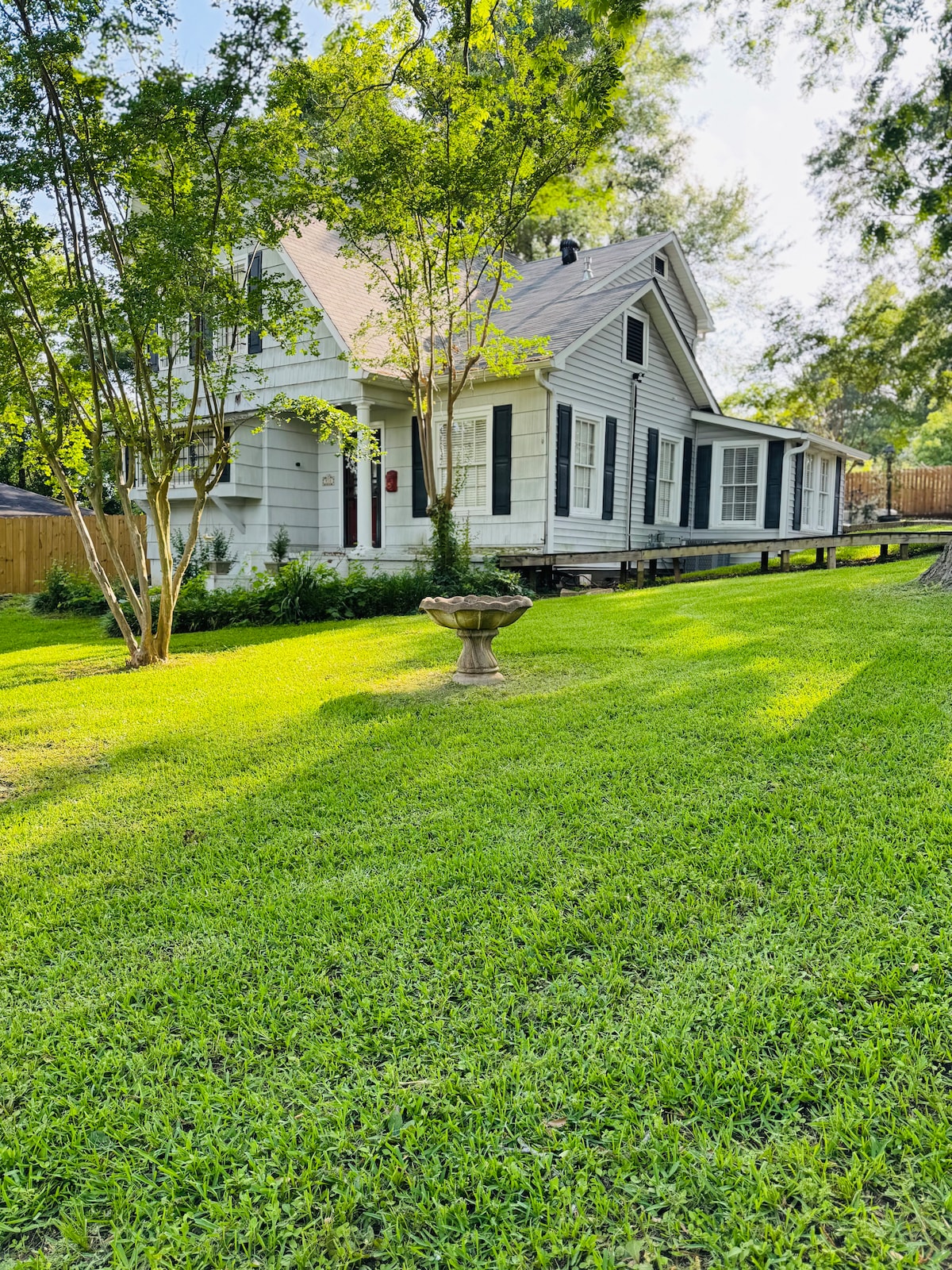 A charming house is framed by lush greenery and well-maintained grass. Tall trees provide shade, while a decorative birdbath stands prominently on the lawn. The structure showcases a blend of classic architecture with a welcoming front porch, enhancing the outdoor space.