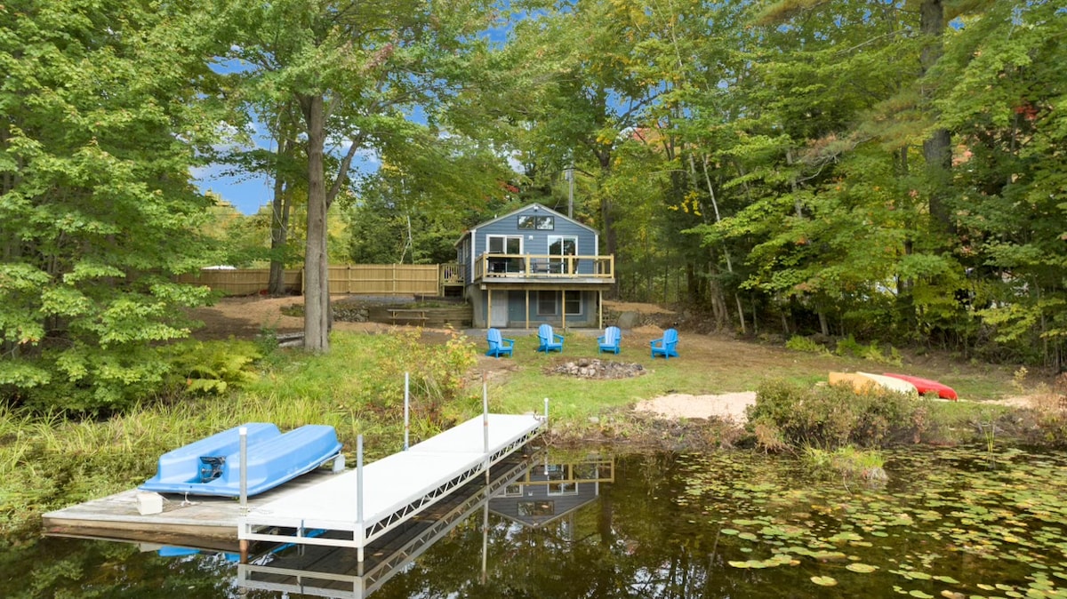 A serene outdoor space features a private dock extending into a pond, surrounded by vibrant greenery. The decked area in front of a modern home includes blue seating and two boats, inviting relaxation and enjoyment by the water.
