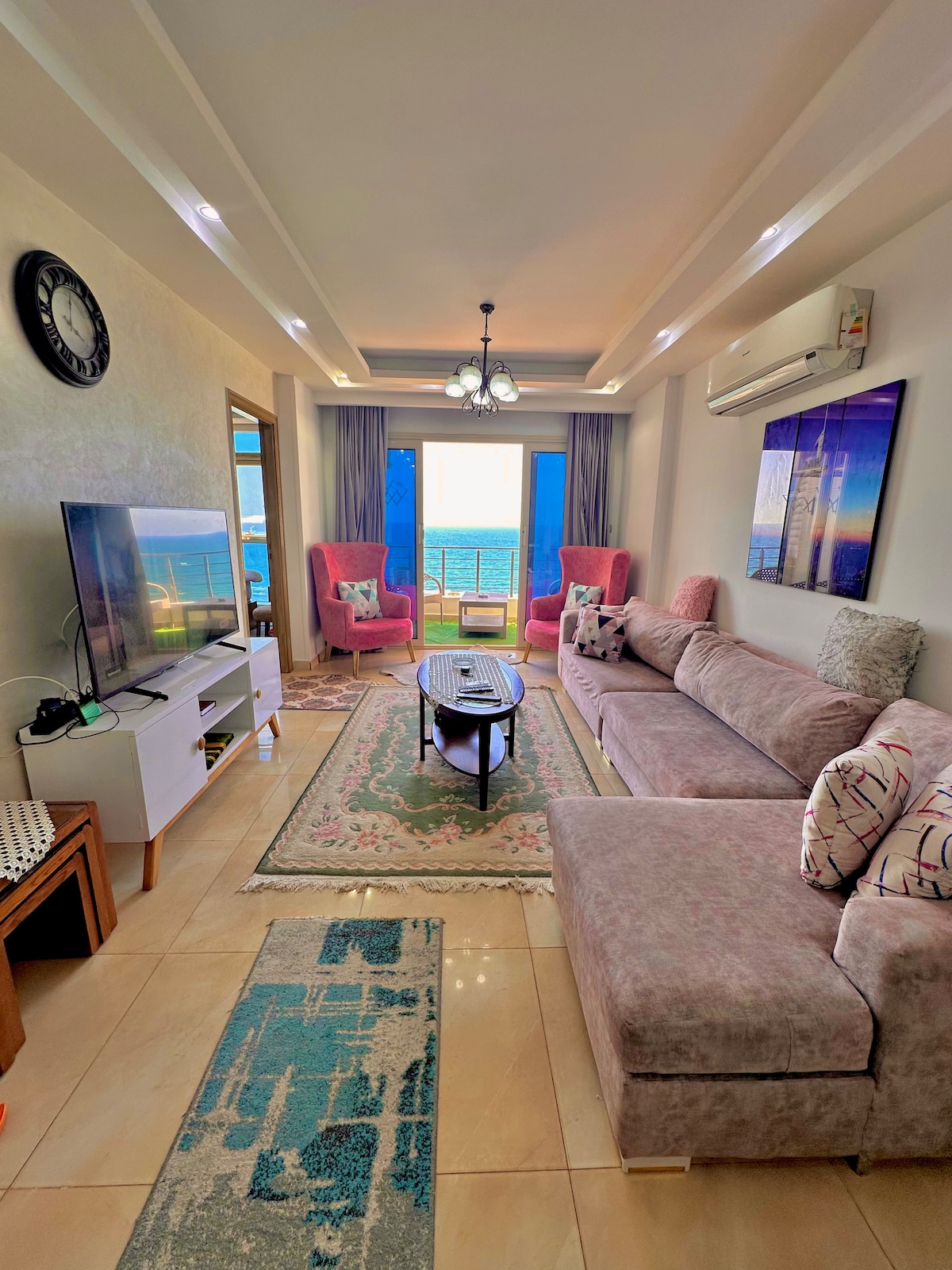 A stylish living room is shown, featuring a plush sectional sofa and two accent chairs facing a coffee table. A large television sits on a white media console. Natural light enters through sliding glass doors, offering views of the sea, while a decorative rug adds warmth to the tiled floor.