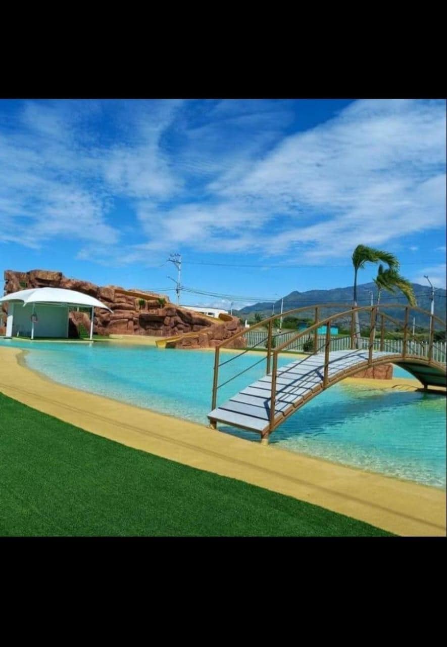 A large, inviting pool area is framed by a bridge leading to a rocky feature in the background. Lush green grass surrounds the pool, and the bright blue sky enhances the tranquil atmosphere.