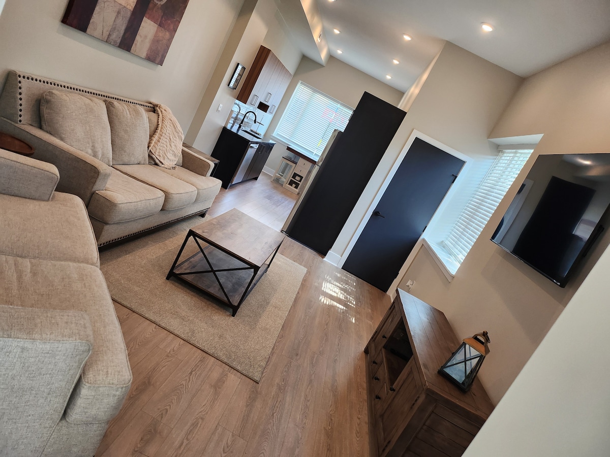 A bright living area is visible, featuring a comfortable beige sofa and a coffee table situated on a light-colored area rug. A mounted television is positioned on the wall opposite a dark door. Natural light enters through the windows, enhancing the spaciousness of the layout.