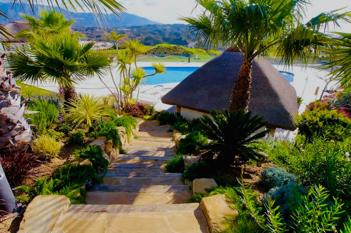 A stone pathway meanders down through lush greenery, flanked by various palm trees and vibrant plants, leading to a pool area in the background. A thatched-roof structure provides shade beside the pool, with scenic hills visible in the distance.