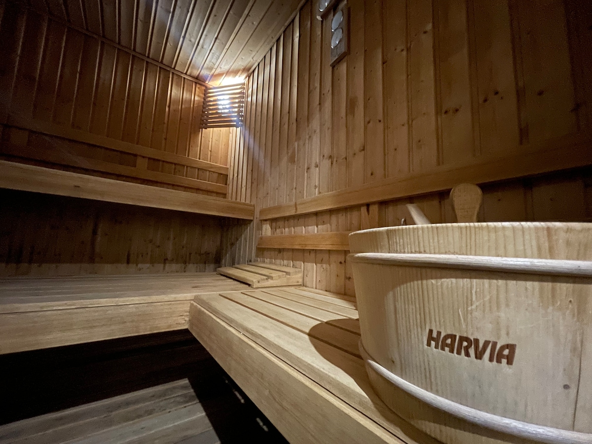 A wooden sauna interior features warm-toned wooden benches and walls. Soft lighting creates a soothing ambiance. A wooden bucket with the branding 'HARVIA' is placed on a bench, adding a practical touch to the tranquil setting.