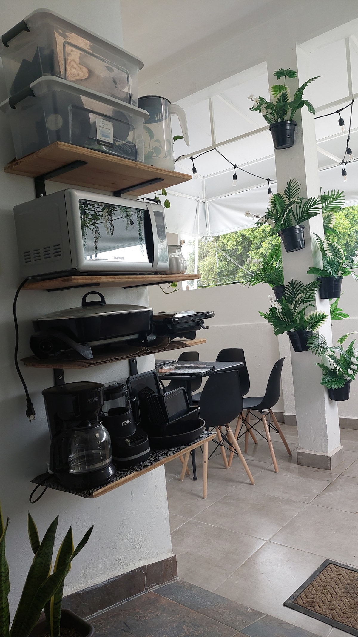 A kitchen area features a tiered shelf holding various appliances, including a microwave and coffee maker. Black dining chairs surround a small table. Green plants are mounted on the walls, complemented by soft lighting from string lights overhead.