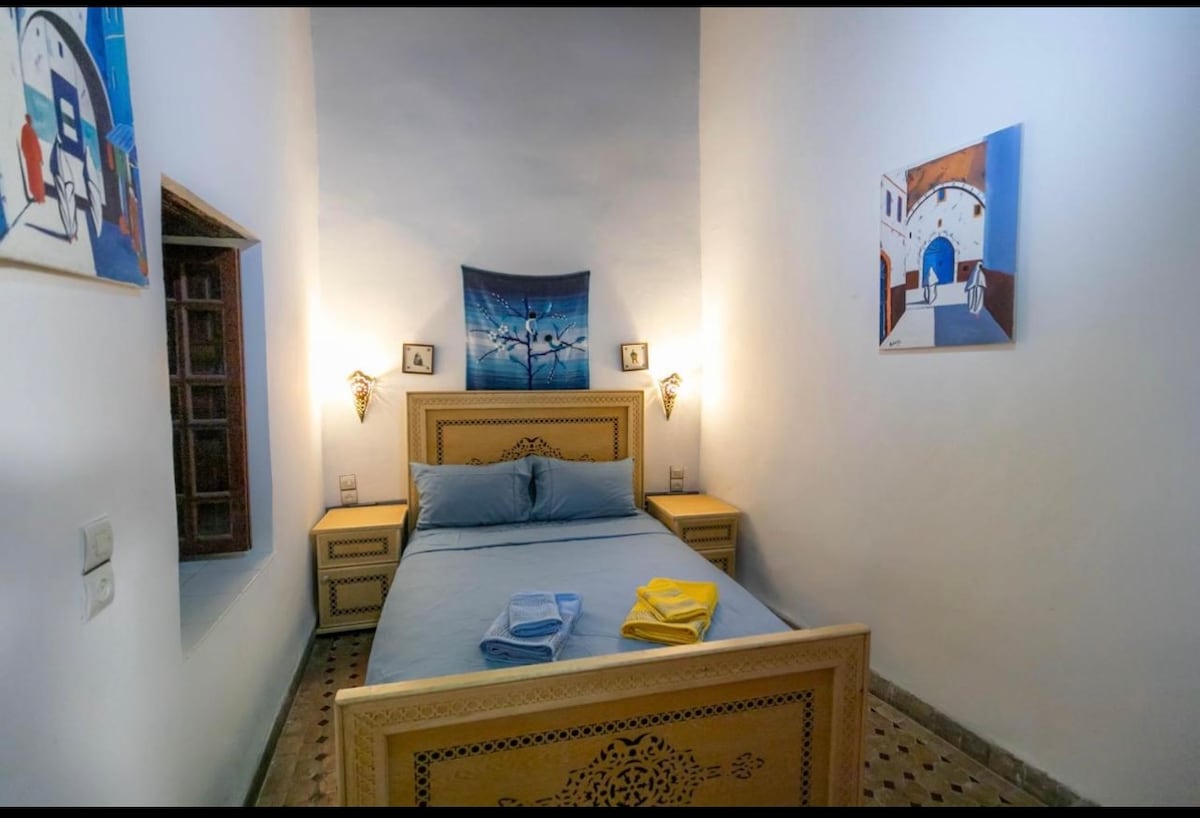 A cozy sleeping area features a wooden bed with a blue duvet and matching pillows. Art pieces adorn the walls, and bedside lamps provide soft lighting. Fresh towels in yellow and blue are neatly placed at the foot of the bed, complementing the minimalist decor.