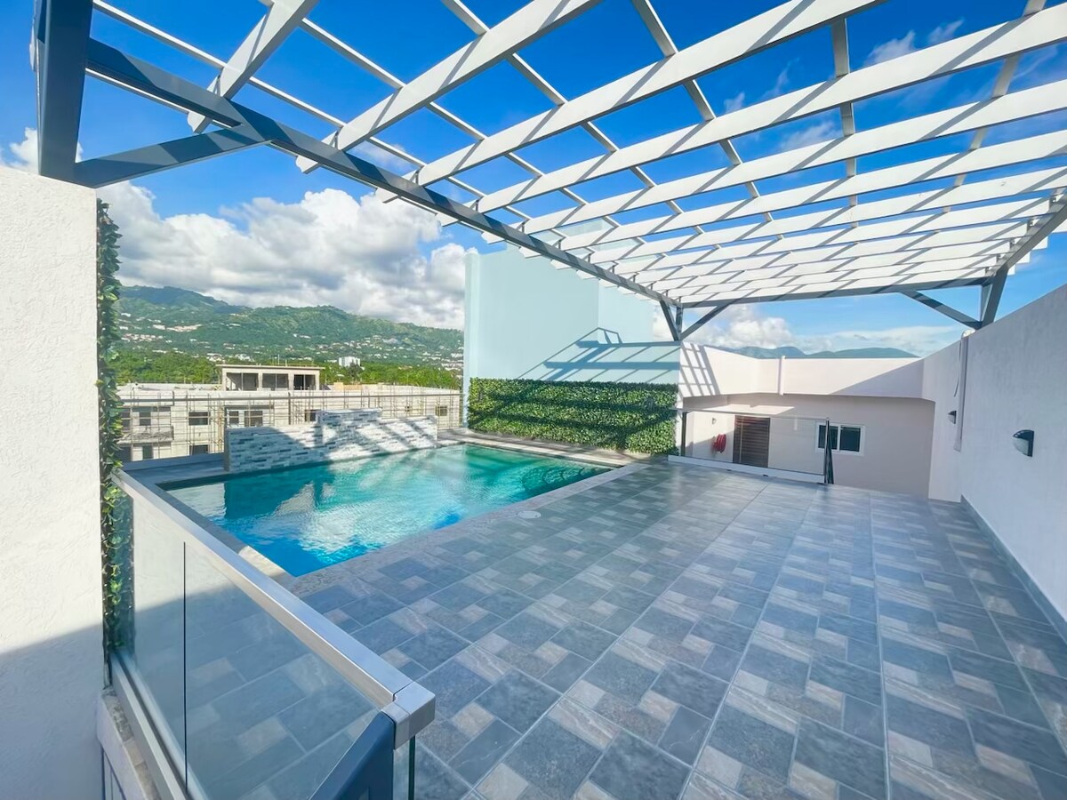 A spacious rooftop area features a large swimming pool surrounded by a stone deck. Natural light filters through a modern pergola, while lush greenery covers the walls, creating a relaxed outdoor retreat with views of the surrounding hills.