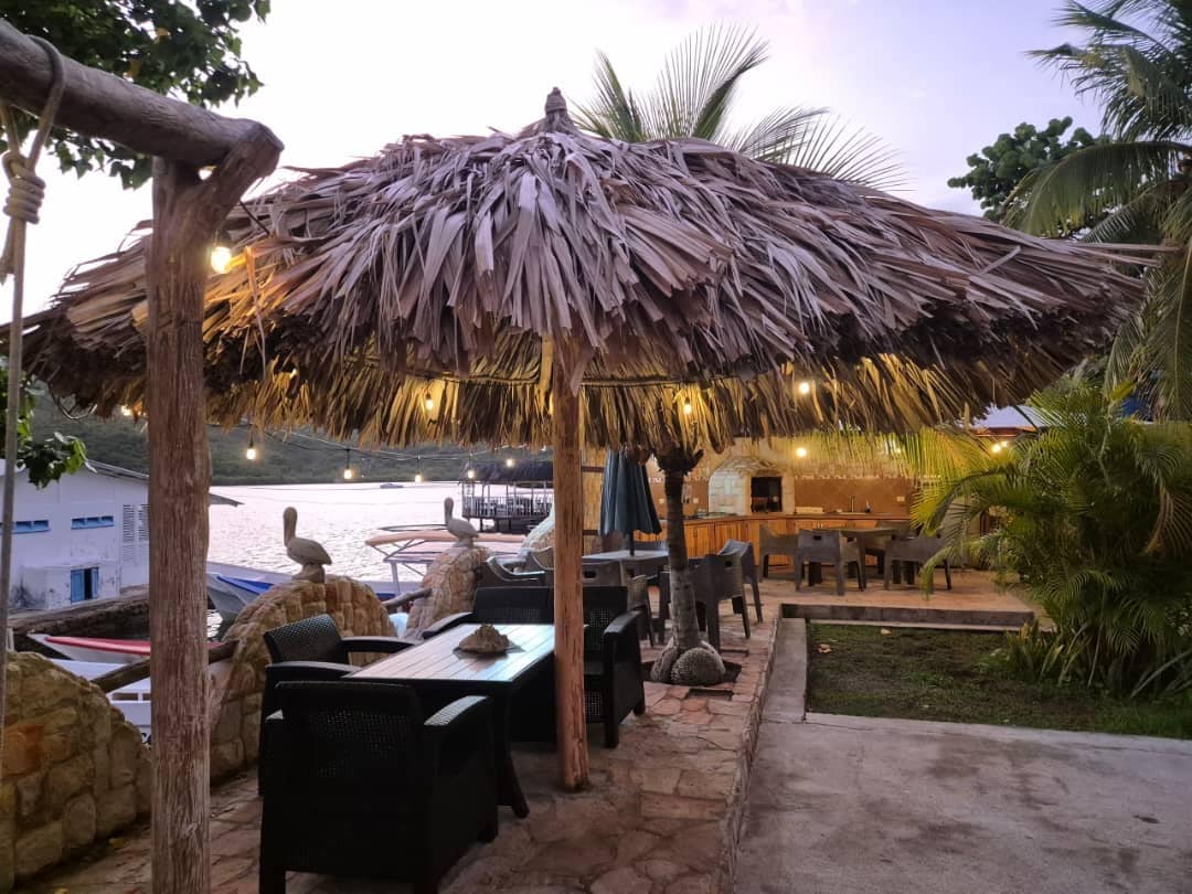 A thatched-roof structure provides shaded seating beneath soft lighting, surrounded by palm trees and stone accents. Tables and chairs are arranged in the outdoor space, with a view of the water visible in the background, enhancing the inviting atmosphere of the area.