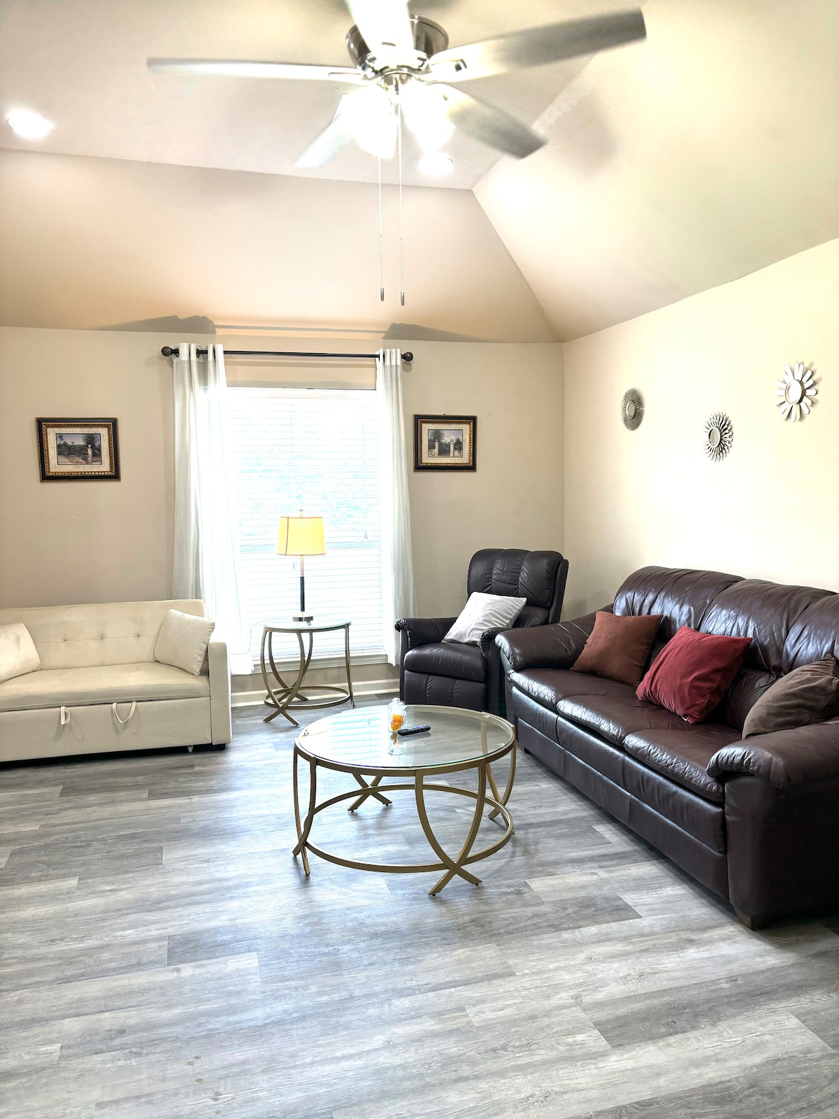 A bright living area is illuminated by natural light streaming through a large window, dressed with sheer white curtains. Comfortable seating includes a dark leather sofa and a light-colored loveseat. A round glass coffee table sits at the center, complemented by decorative wall art.