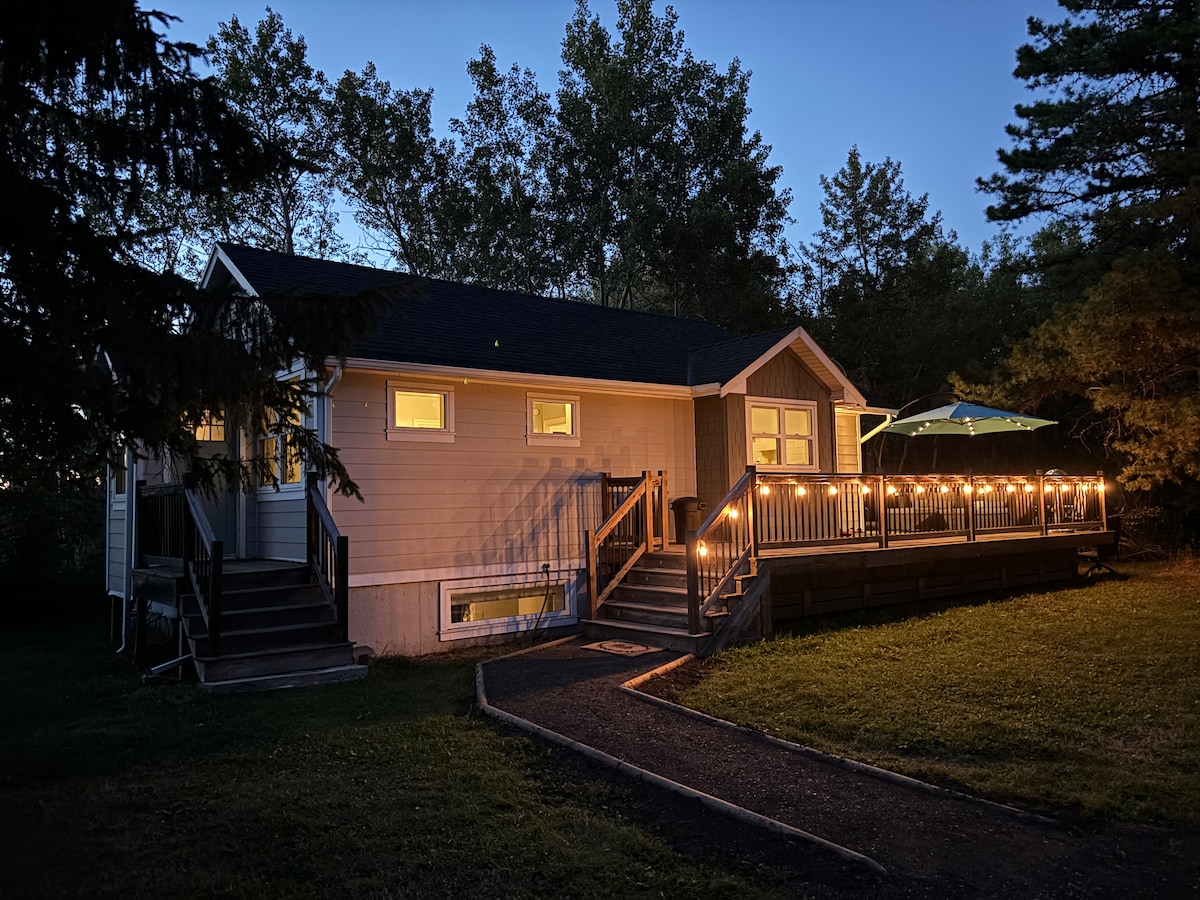 The home is shown at dusk, featuring a spacious deck with string lights illuminating the area. A green umbrella offers shade over part of the deck, while the soft glow from the windows highlights the renovated exterior.