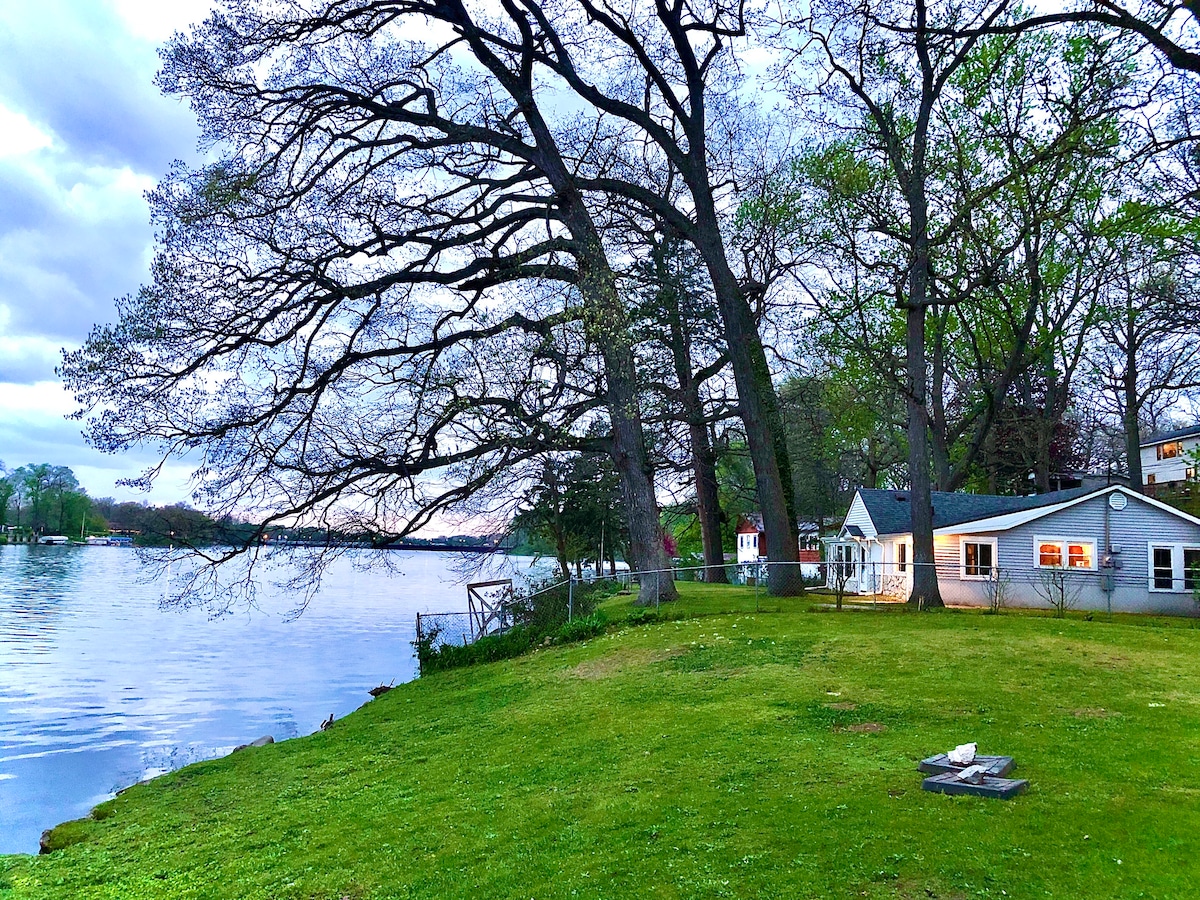 A spacious lawn leads to a serene riverfront, framed by tall trees. A charming cottage is positioned nearby, with soft lighting illuminating its exterior. The water reflects the evening sky, creating a peaceful ambiance. A fire pit is visible on the grassy area.