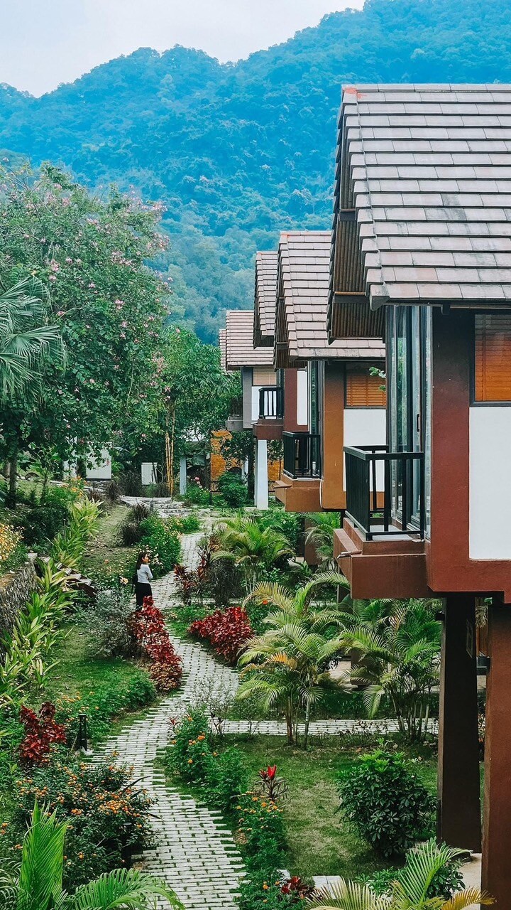 Charming accommodation units are nestled among lush greenery, with an inviting pathway lined with vibrant flowers leading through the garden. Mountains provide a scenic backdrop, enhancing the tranquil atmosphere of the resort.