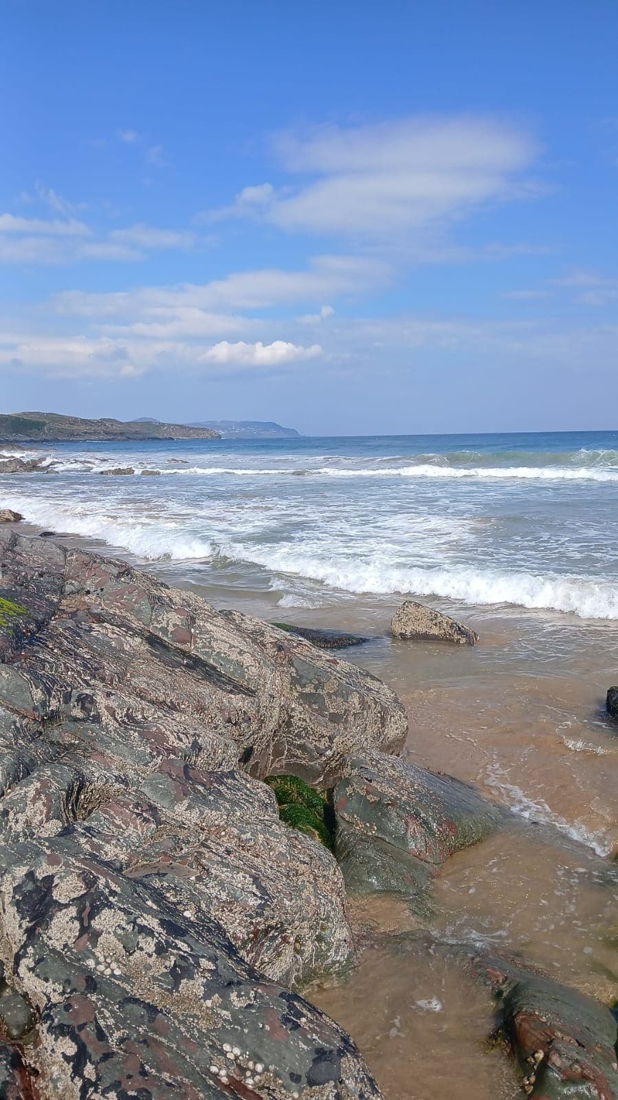 The image showcases a serene coastline where gentle waves meet rugged, textured rocks. A clear blue sky filled with soft clouds complements the calm waters, reflecting shades of blue and white. The sandy beach is partially visible, illustrating a peaceful seascape.