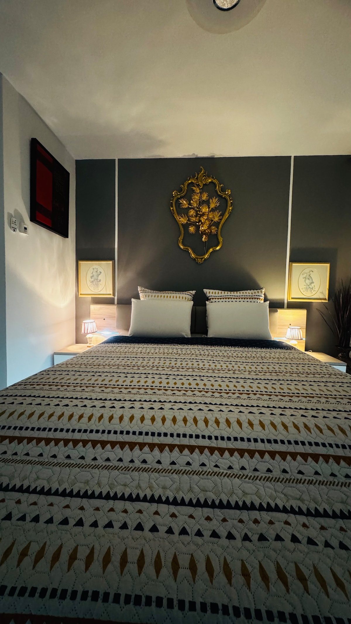 A well-appointed sleeping area features a spacious bed with decorative pillows and an intricately patterned bedspread. Soft lighting from two nightstands enhances the room's ambiance. A decorative wall piece hangs above the bed against a neutral backdrop, contributing to a serene environment.
