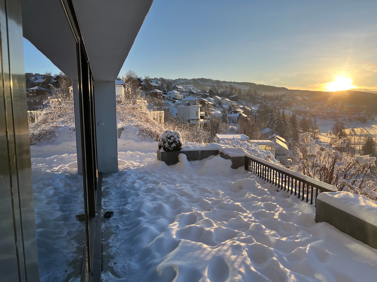A spacious outdoor terrace is shown, blanketed in fresh snow, with a panoramic view of the surrounding hills and houses. The setting sun casts a warm glow over the landscape, enhancing the scenic winter atmosphere.