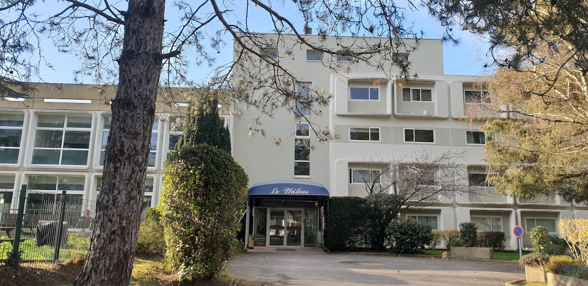 The entrance of a residential hotel is shown, featuring a covered awning labeled 'Le Hôtelen.' Large windows are visible on the building's façade, surrounded by lush greenery and trees. A pathway leads to the entrance, blending the structure with its natural surroundings.
