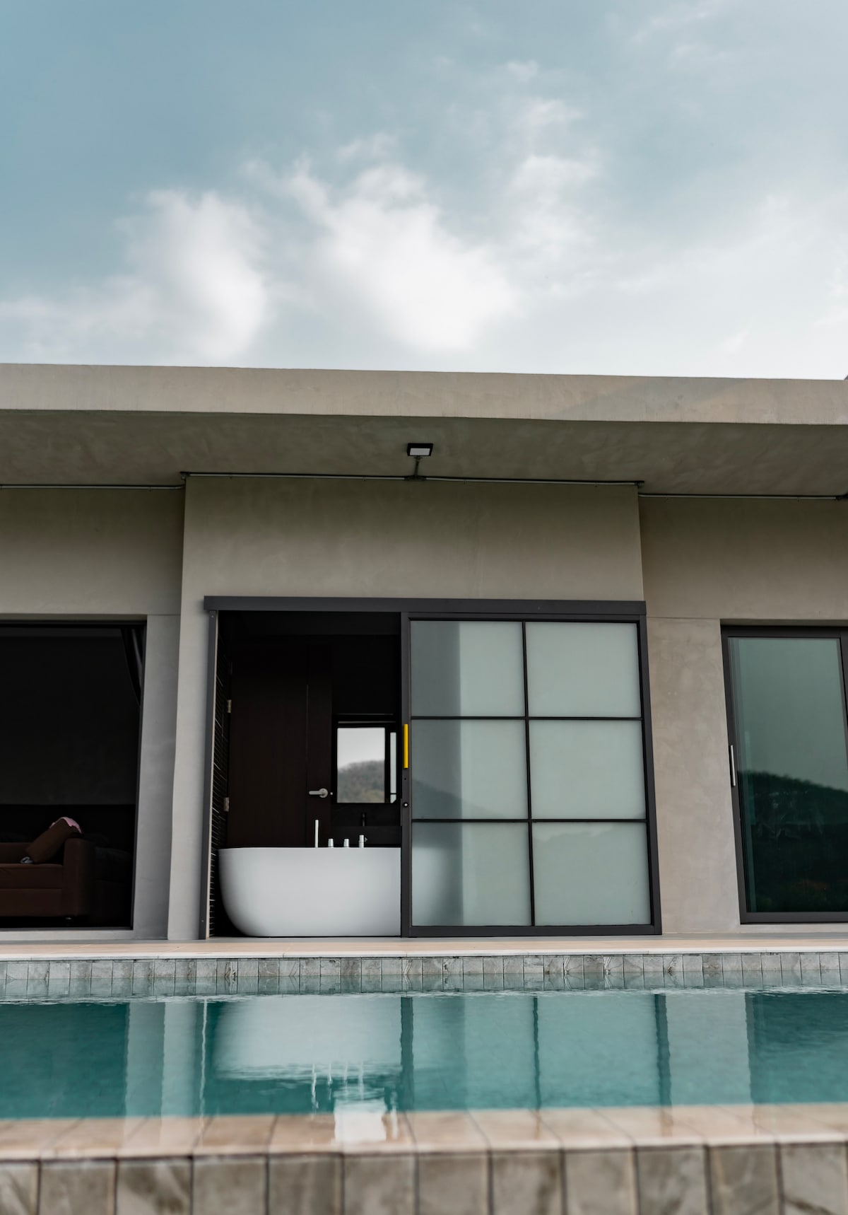 A modern villa is depicted with a spacious outdoor area featuring a clear swimming pool. Large glass doors provide a view of an indoor bathtub, while neutral-colored walls and a minimalist design enhance the overall tranquility of the setting.