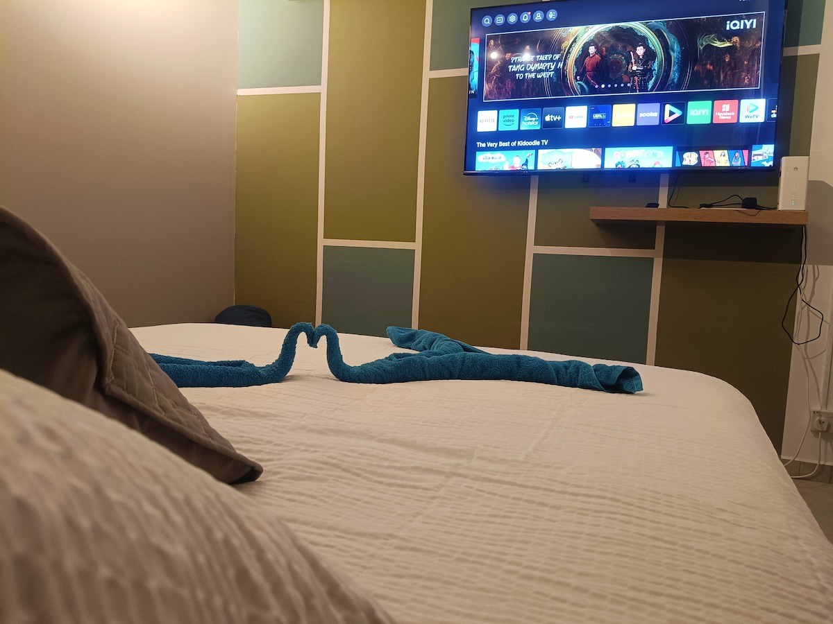 A spacious bed is centered in the room, featuring crisp white linens and a neatly arranged blue towel. A large smart TV is mounted on the wall above a shelf, displaying various streaming options against a backdrop of geometric wall panels in muted colors.