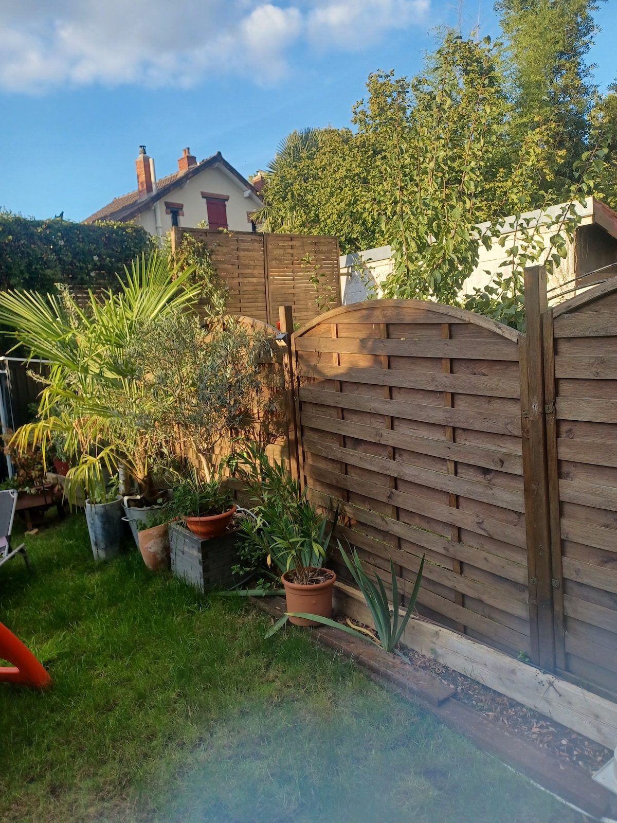 A small garden area is surrounded by wooden fencing, featuring a variety of potted plants and greenery, including palm and aloe plants. The lawn is well-maintained, providing a natural setting. Bright sunlight filters through the foliage, creating a peaceful outdoor space.