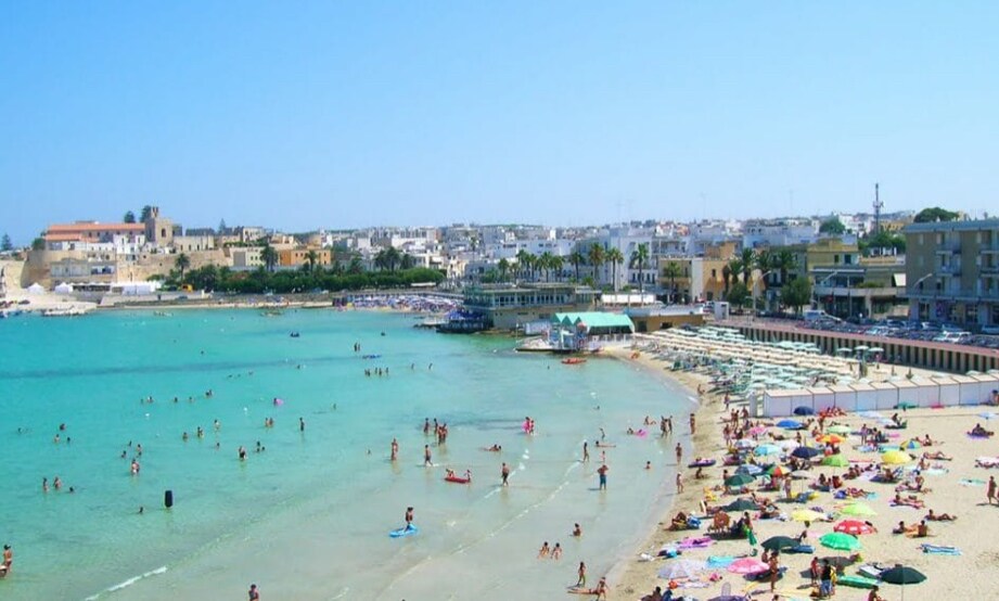 A panoramic view captures a vibrant beach area with turquoise waters lapping at a sandy shore. Sunbathers are seen on colorful towels and beach umbrellas, while light waves create a gentle shoreline. In the background, buildings and coastal structures are visible along the promenade.