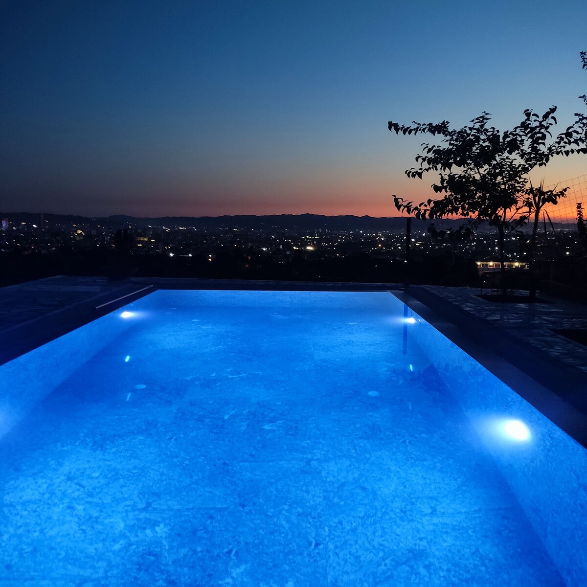 A serene pool glows with blue lighting, set against a backdrop of a dusky sky. Silhouettes of trees frame the edge, enhancing the tranquil atmosphere of the outdoor space. The distant horizon shows gentle hills lit by city lights in the twilight.