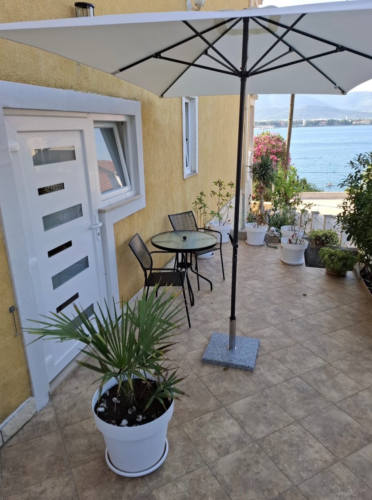 A spacious terrace area is visible, featuring a glass table with two black chairs under a white umbrella. Potted plants surround the space, and a view of the sea is in the background against a yellow exterior wall.
