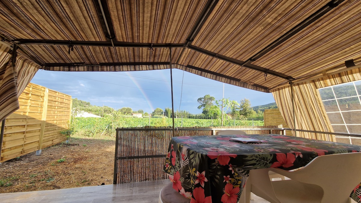 A shaded terrace is presented with a patterned tablecloth covering a round table. Nearby, a rainbow arcs over lush greenery visible beyond wooden fencing. Natural light fills the area, highlighting the open space and inviting outdoor experience.