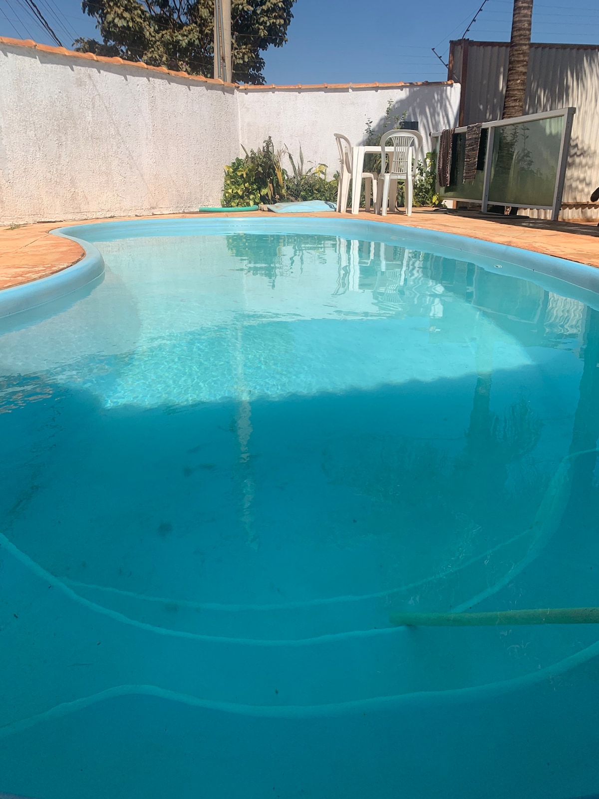 A clear swimming pool is visible, reflecting sunlight and surrounded by a well-maintained patio. The area features greenery along the pool's edge, with a small table and chairs positioned nearby for relaxation. A section of a building can be seen in the background.