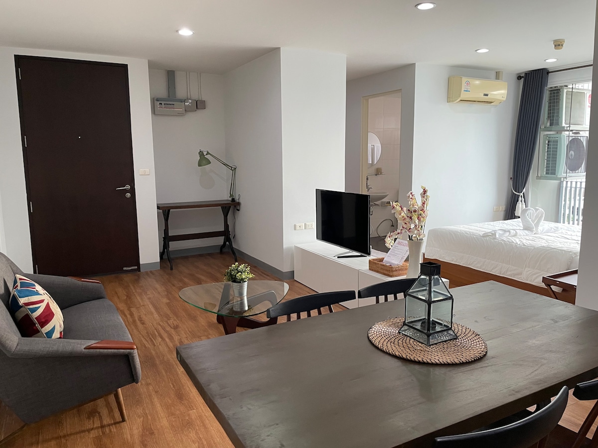 A modern studio apartment features a spacious layout with wood flooring. A comfortable seating area includes a gray armchair and a small glass table. A television is positioned on a white unit, while the sleeping area is visible, showcasing a neatly made bed. Natural light is provided by large windows.