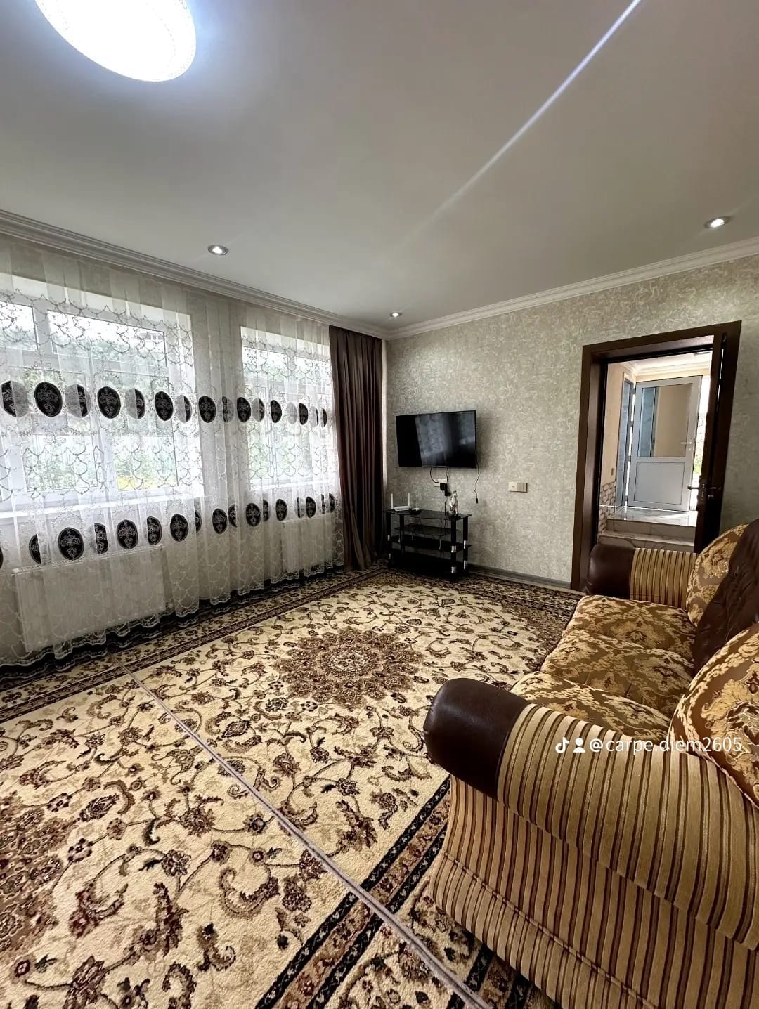 An inviting living area features a patterned area rug on a wooden floor. Large windows are adorned with sheer curtains, allowing natural light to fill the space. A flat-screen TV is mounted on the wall, and a comfortable sofa offers a place for relaxation.