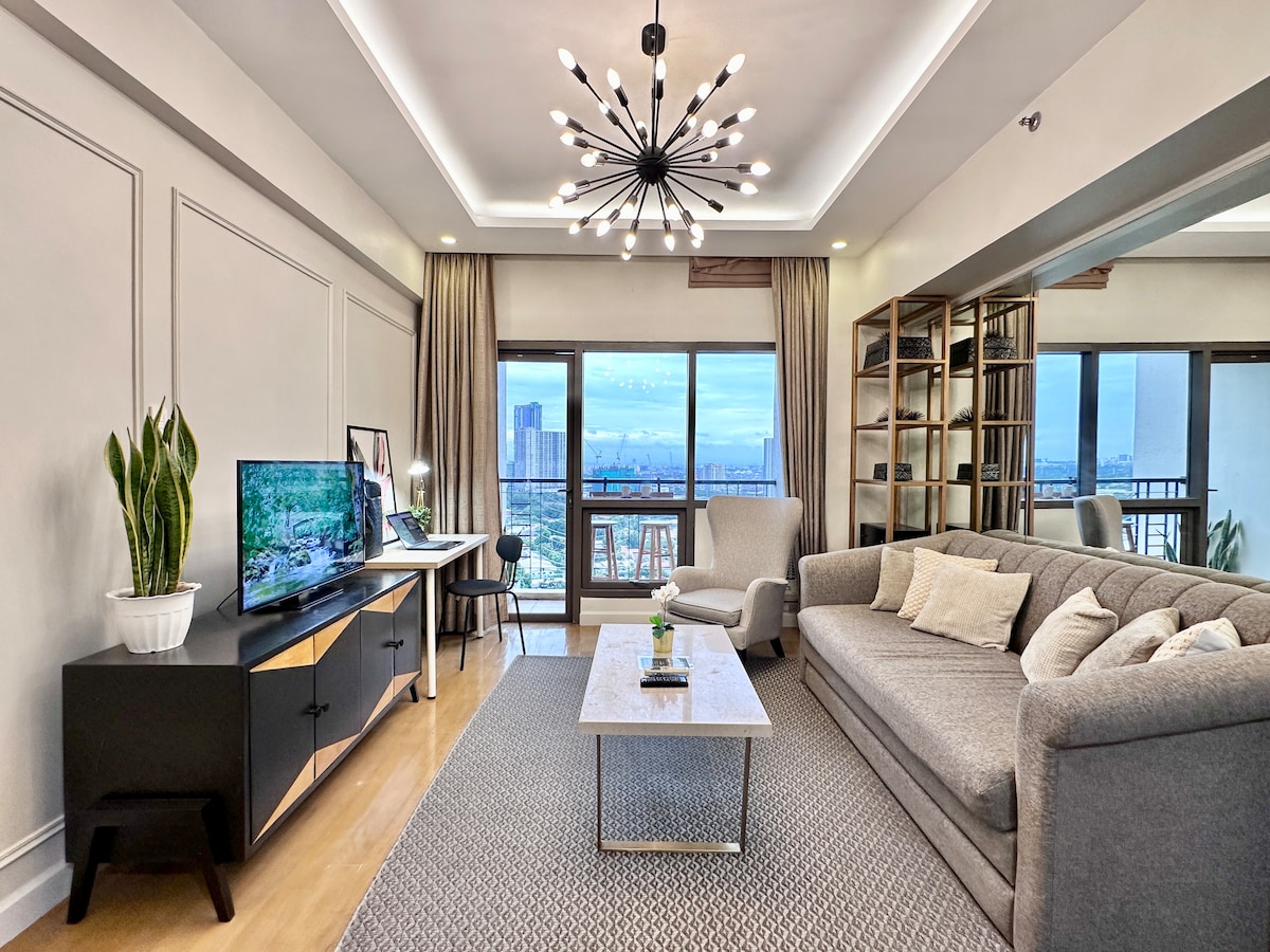 A comfortable living area is presented with a three-seater sofa bed, a coffee table, and a large TV mounted on a media console. A live plant is visible, and large windows open to views of the city, while modern lighting features above enhance the space.