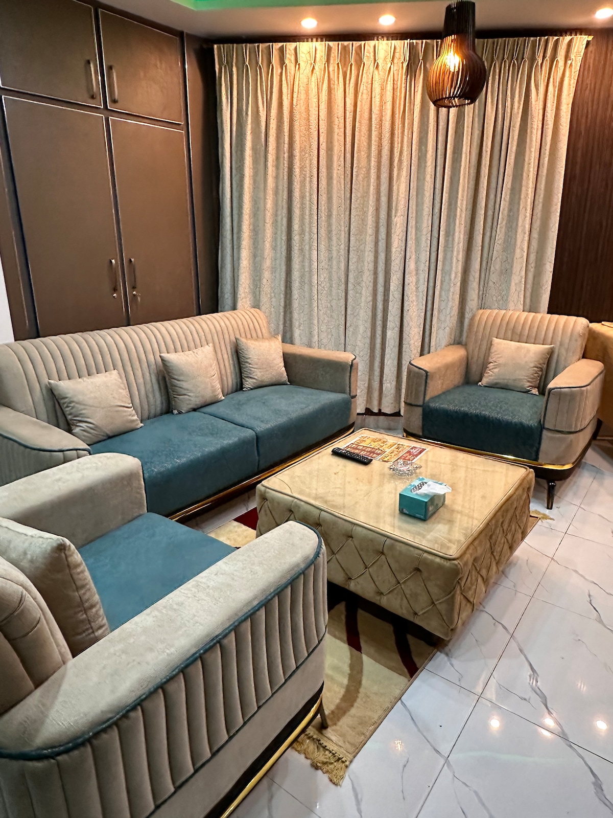 A cozy drawing room features a seating arrangement of two armchairs and a matching sofa set, all with soft fabric. A low coffee table sits at the center, surrounded by patterned rugs. Light filters in through sheer curtains, adding to the calm ambiance.