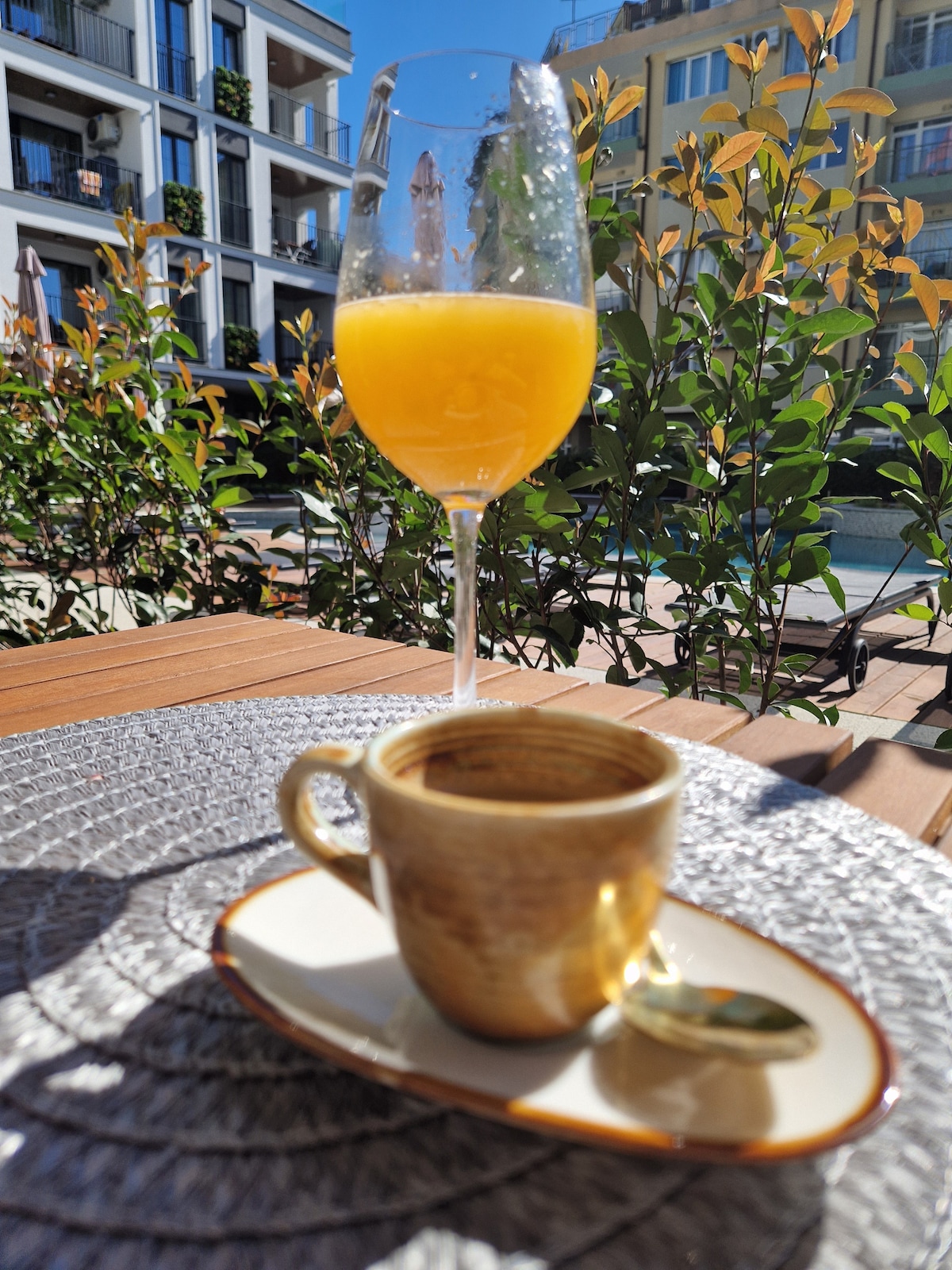 A refreshing glass of orange juice is positioned beside a small, intricately designed espresso cup on a textured table. Soft greenery and hints of a pool area are visible in the background, illuminated by bright sunlight.