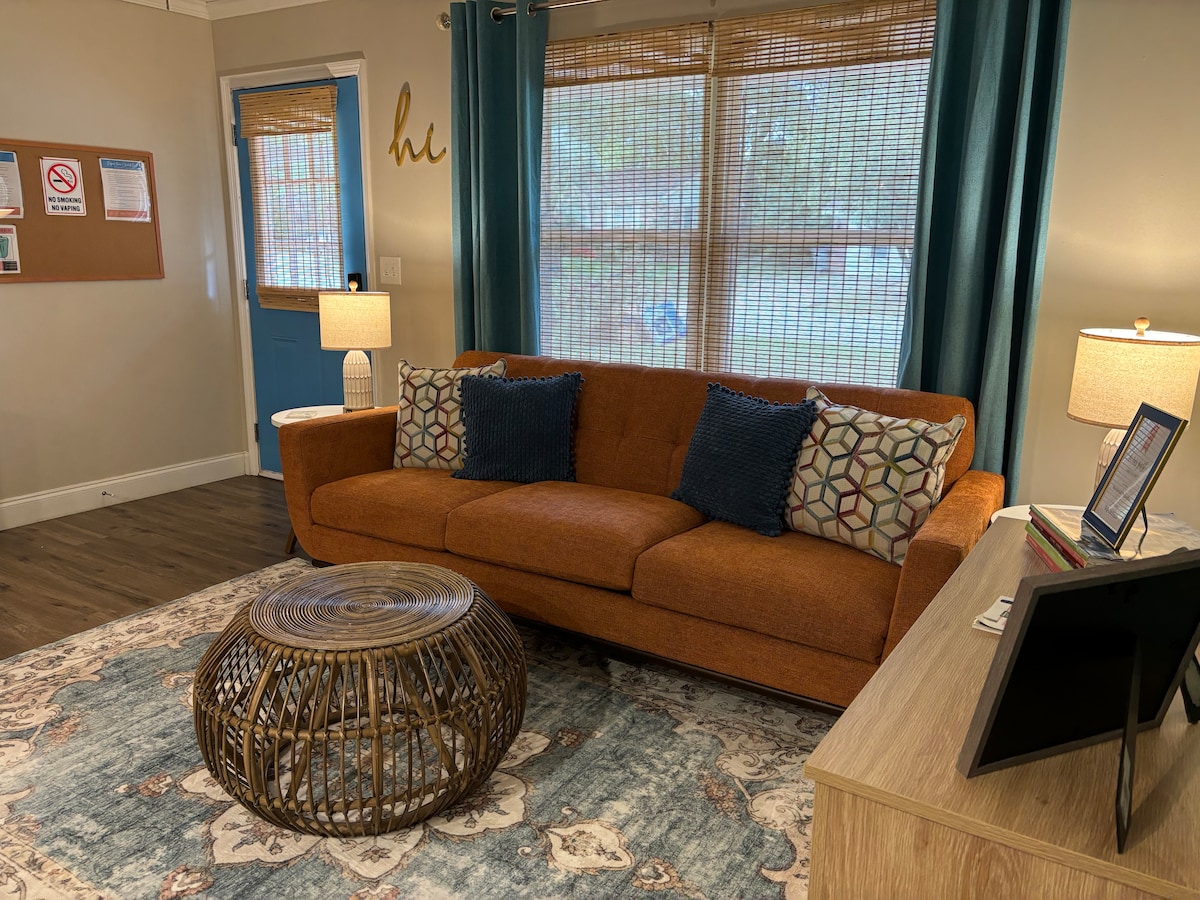 A warm living area features a comfortable orange sofa adorned with patterned cushions. A round woven coffee table sits in front, complemented by a light-colored area rug. Soft light is provided by two bedside lamps, with natural light filtering through the window covered by bamboo shades.