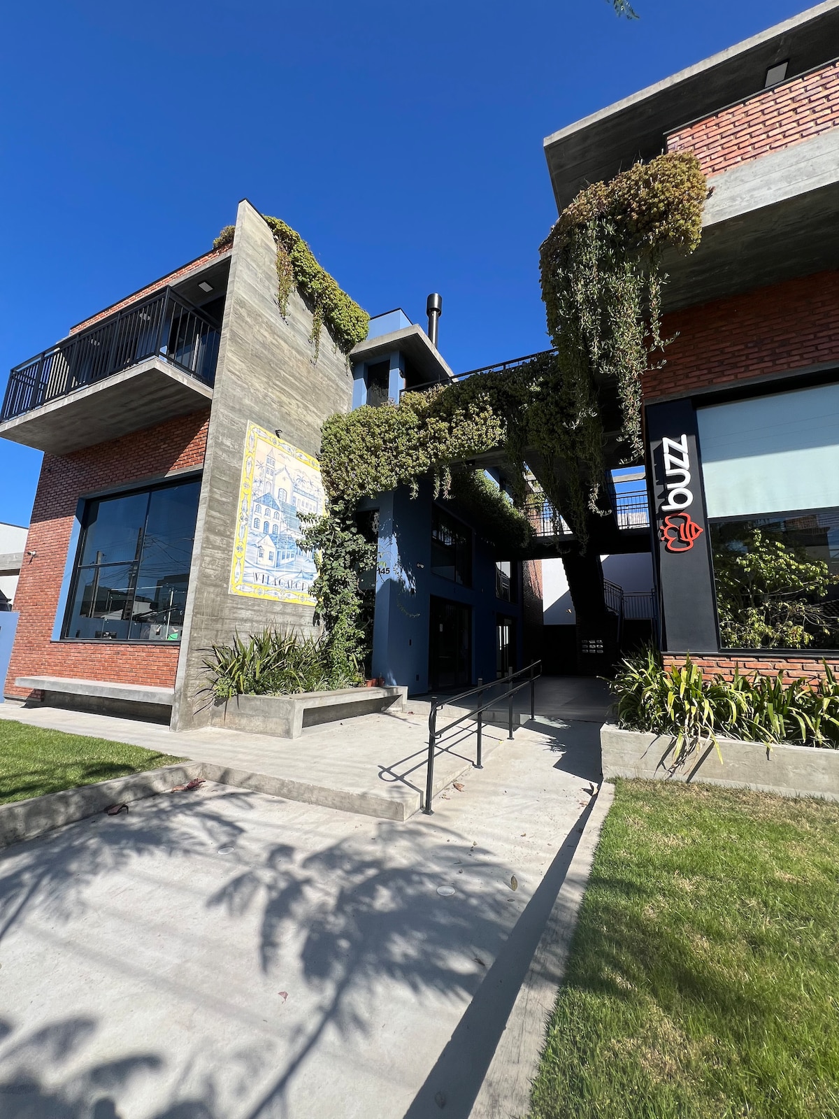 A modern multi-level building features a combination of brick and concrete walls, with greenery cascading over the exterior. A wide walkway leads to the entrance, which is accented by large windows and a stylish sign. The clear blue sky adds to the contemporary charm.