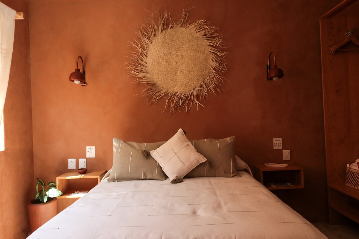 A spacious bed features neutral-toned linens and decorative pillows, positioned against a warm clay wall. Wall-mounted lamps provide soft lighting alongside natural decor, including a woven wall hanging. Nightstands on either side offer functional space, enhancing a cozy atmosphere.