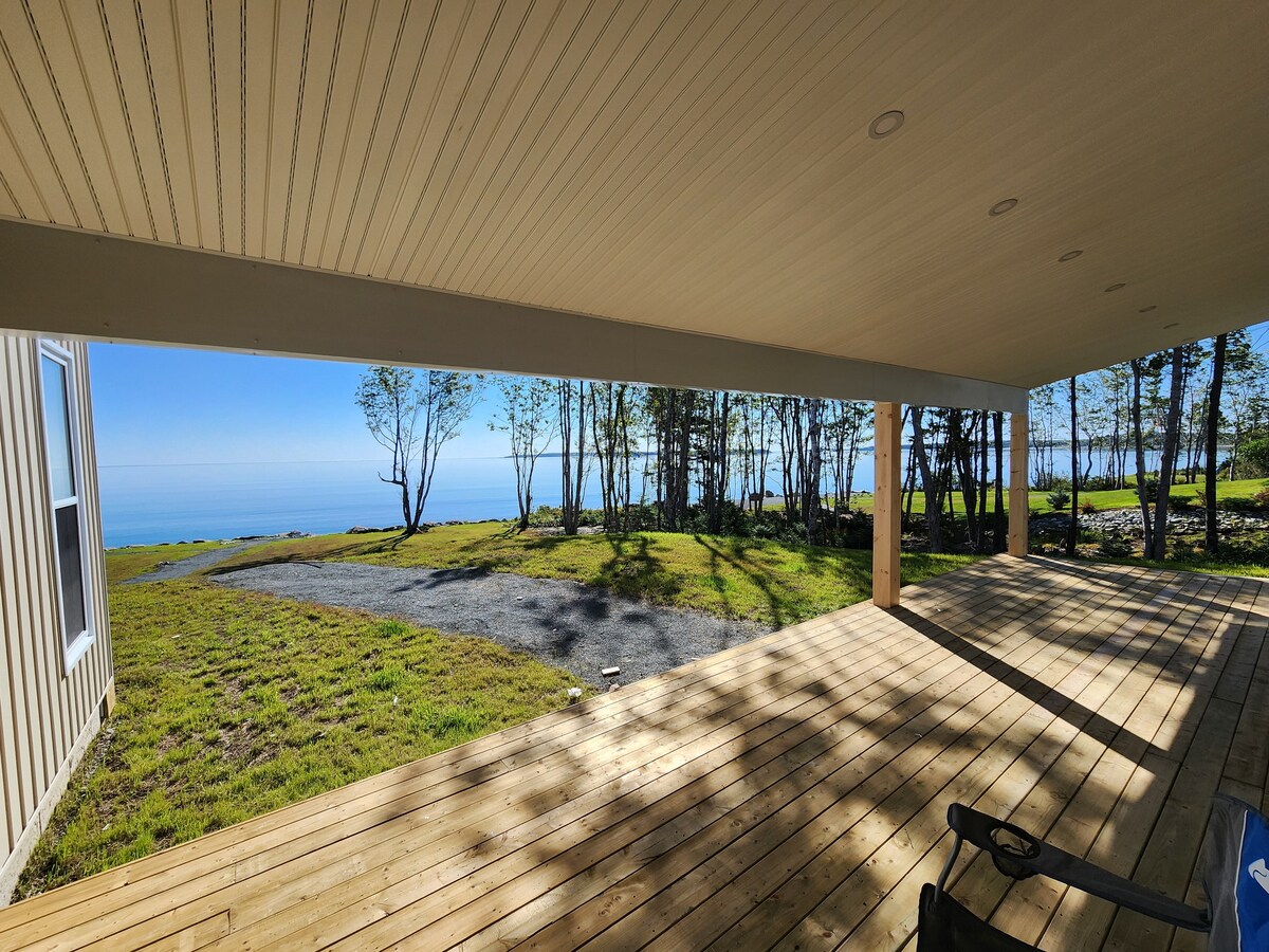A covered outdoor area showcases a spacious wooden deck, extending towards a grassy patch. The Atlantic Ocean can be seen in the distance, framed by trees and blue sky. Natural light filters through, highlighting the tranquil surroundings.