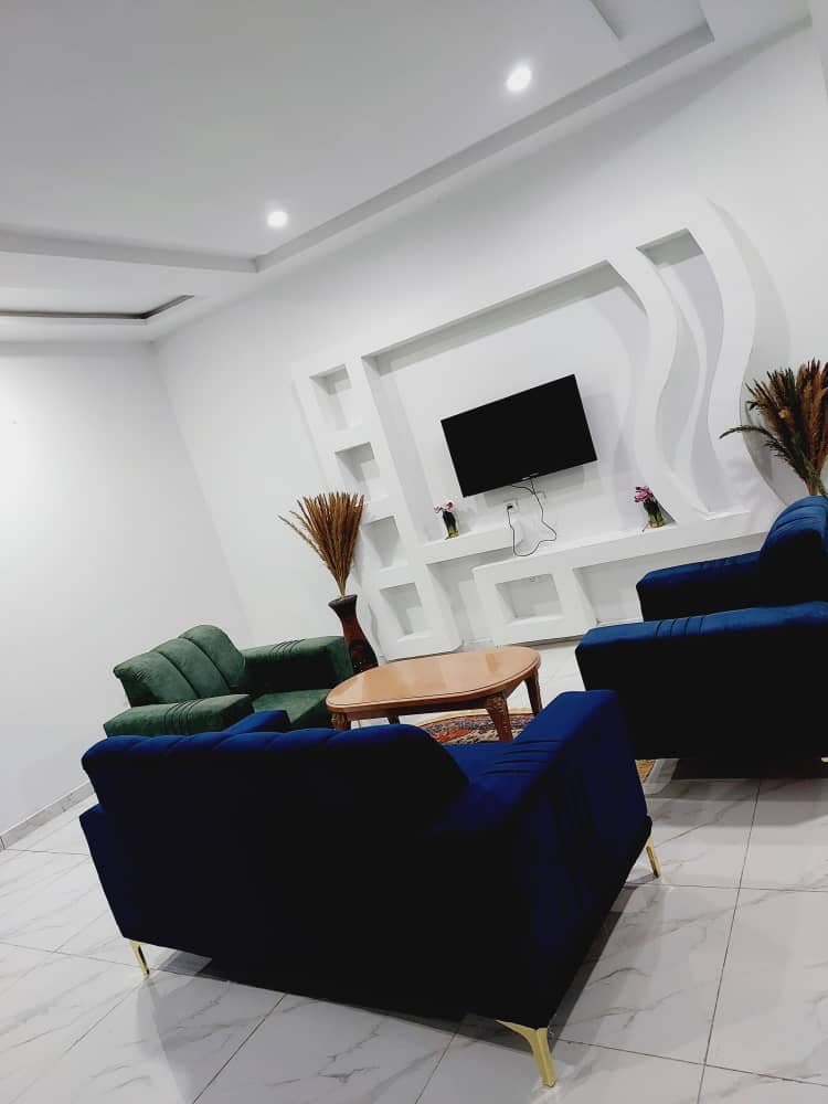 A contemporary living area is presented with a light-colored floor and white walls. Two dark blue sofas and a green loveseat surround a small wooden coffee table. A wall-mounted television is situated above a stylish display niche, complemented by decorative plants in the corners.