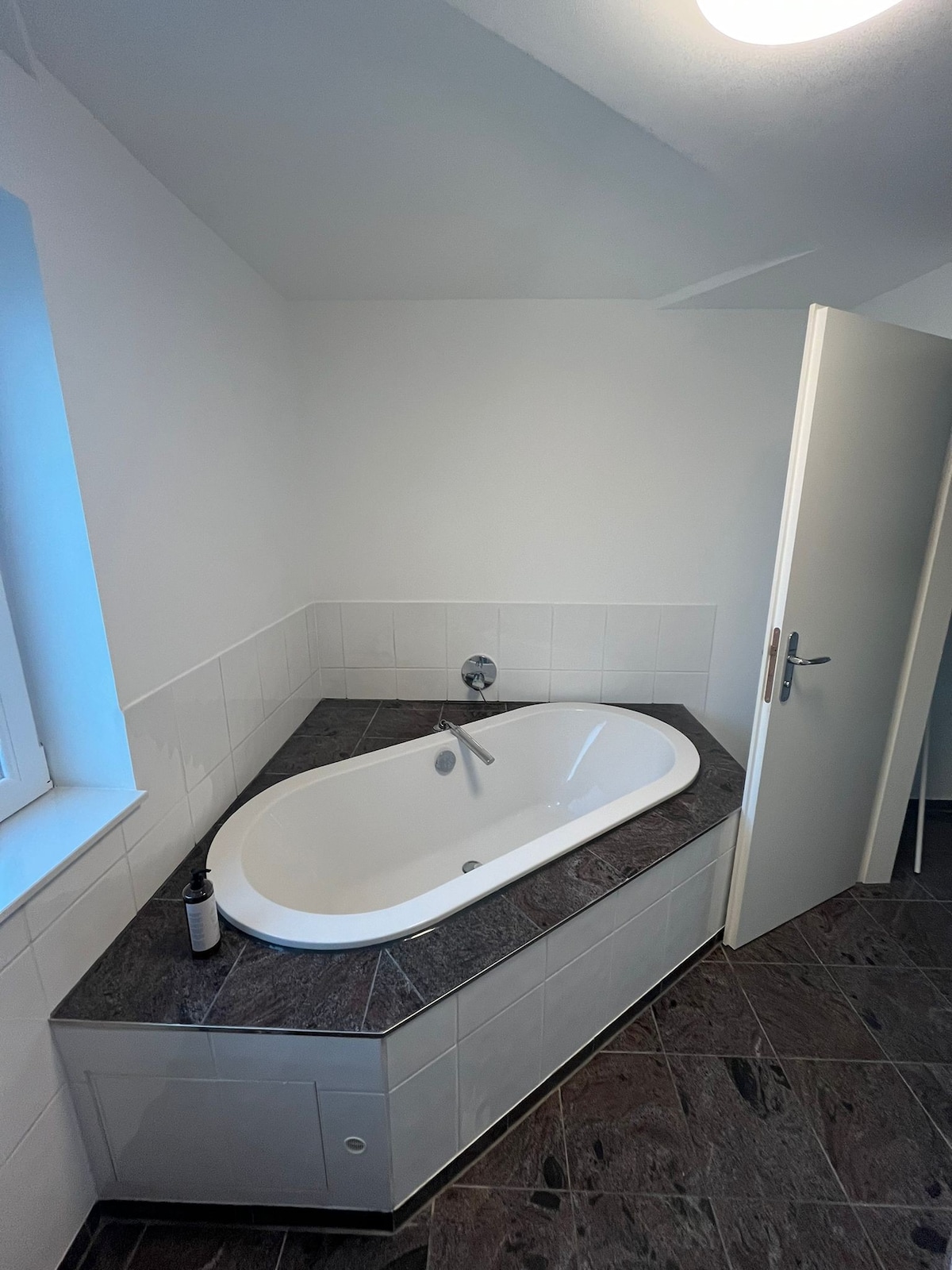 A modern bathroom features an elegant corner bathtub set on a dark stone surface. White tiles cover the walls, and a door leads to another area. Natural light enters through a nearby window, enhancing the spacious feel of the room.