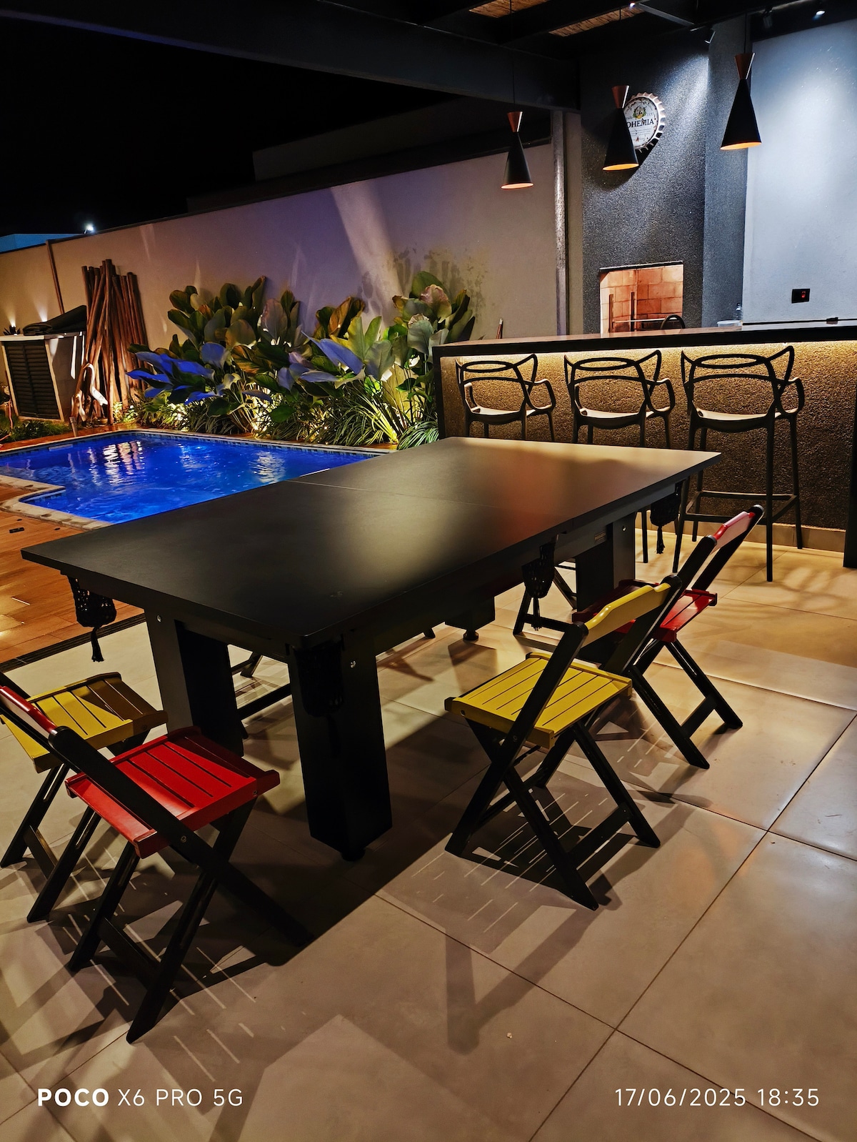 An outdoor area features a modern table surrounded by colorful folding chairs. In the background, a pool is illuminated, complementing the sleek aesthetic. Plants add greenery, while the bar area is visible, creating an inviting space for gatherings.