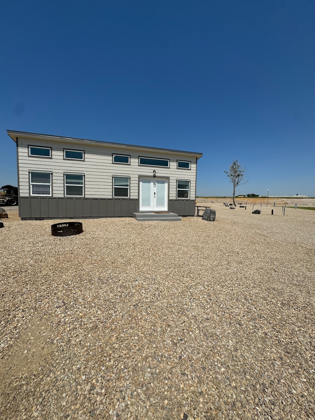 The exterior of a modern tiny home is presented, featuring a clean design with multiple large windows and a welcoming front door. A gravel area surrounds the house, with open space visible in the background, enhancing the serene setting.