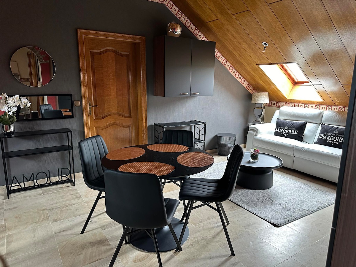 An inviting living area features a circular dining table surrounded by four black chairs. A cozy sofa is positioned against the wall, complemented by neutral decor. Natural light enters through a skylight, illuminating the space, while a tasteful arrangement of plants and decorative items adds character.