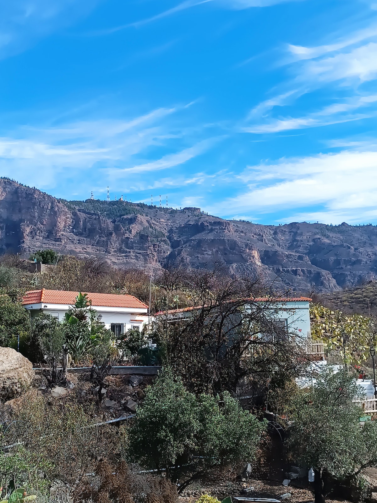 A rustic property is nestled against a mountainous backdrop, featuring weathered trees and shrubs in the foreground. The structure displays a sloped roof with terracotta tiles, surrounded by a rocky landscape under a bright blue sky adorned with wispy clouds.