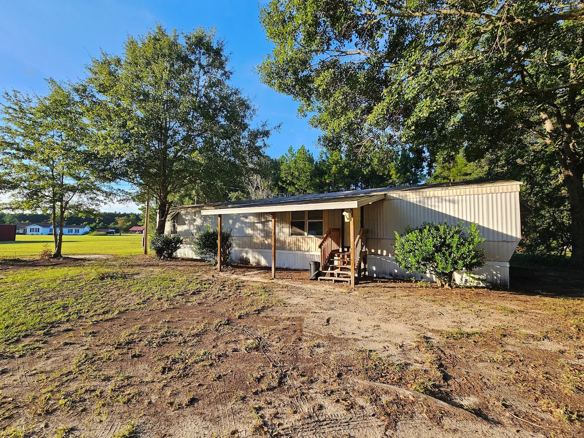 A single-wide manufactured home is situated on a spacious grassy lot, surrounded by trees. A wooden front porch with steps leads up to the entrance. The area is marked by a tranquil rural atmosphere, providing ample outdoor space for relaxation.