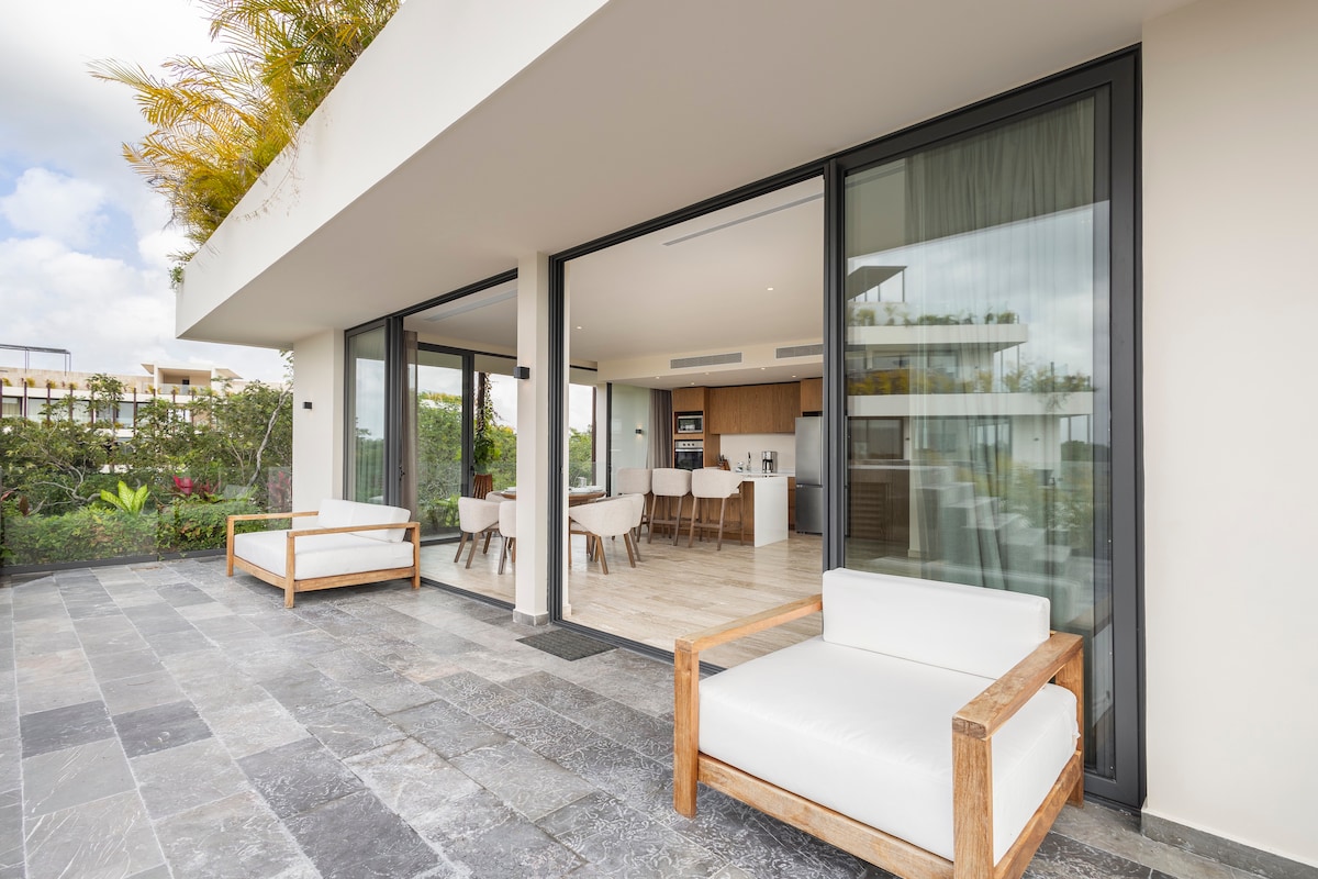 An outdoor terrace is showcased, featuring two modern white lounge chairs framed with natural wood. Floor-to-ceiling glass doors connect to a sunlit interior, highlighting a spacious living area and a dining setup visible through the glass. Lush greenery surrounds the space.