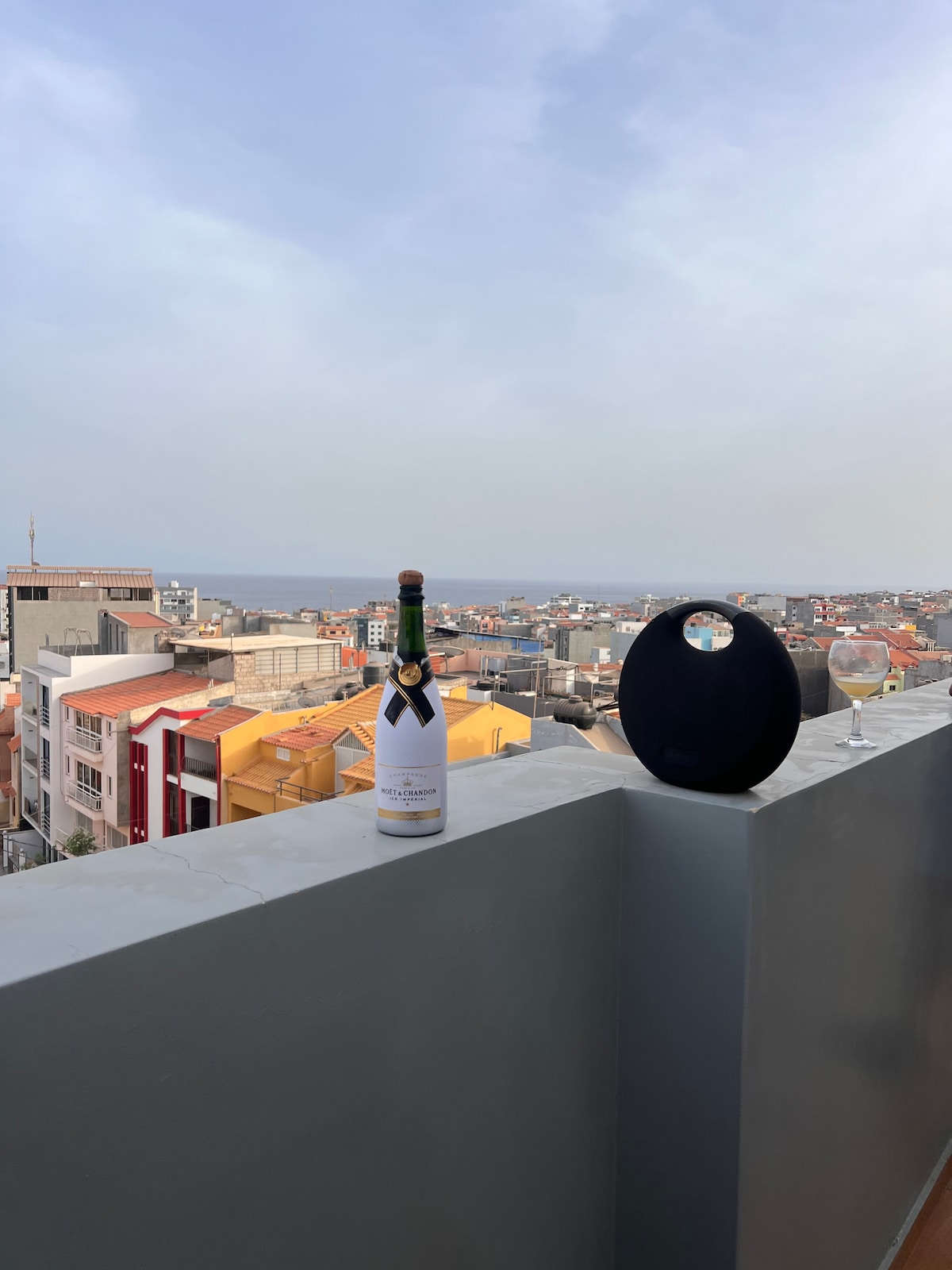An outdoor balcony overlooks a vibrant cityscape. A bottle of champagne stands next to an empty glass and a round speaker on a smooth ledge. The skyline features an array of colorful buildings and distant rooftops under a cloudy sky.