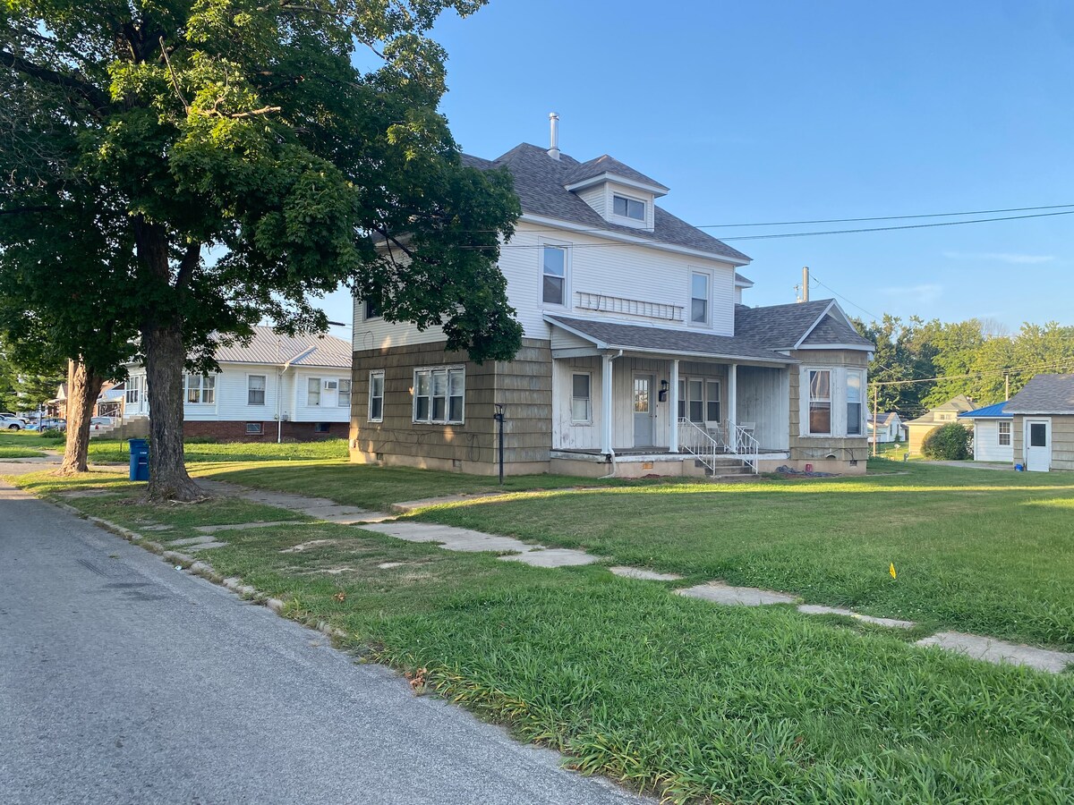 An exterior view of a two-story apartment building is showcased, featuring a combination of light siding and stone accents. A spacious porch with steps leads to the entrance. Lush green grass borders the sidewalk, while trees provide shade along the street.