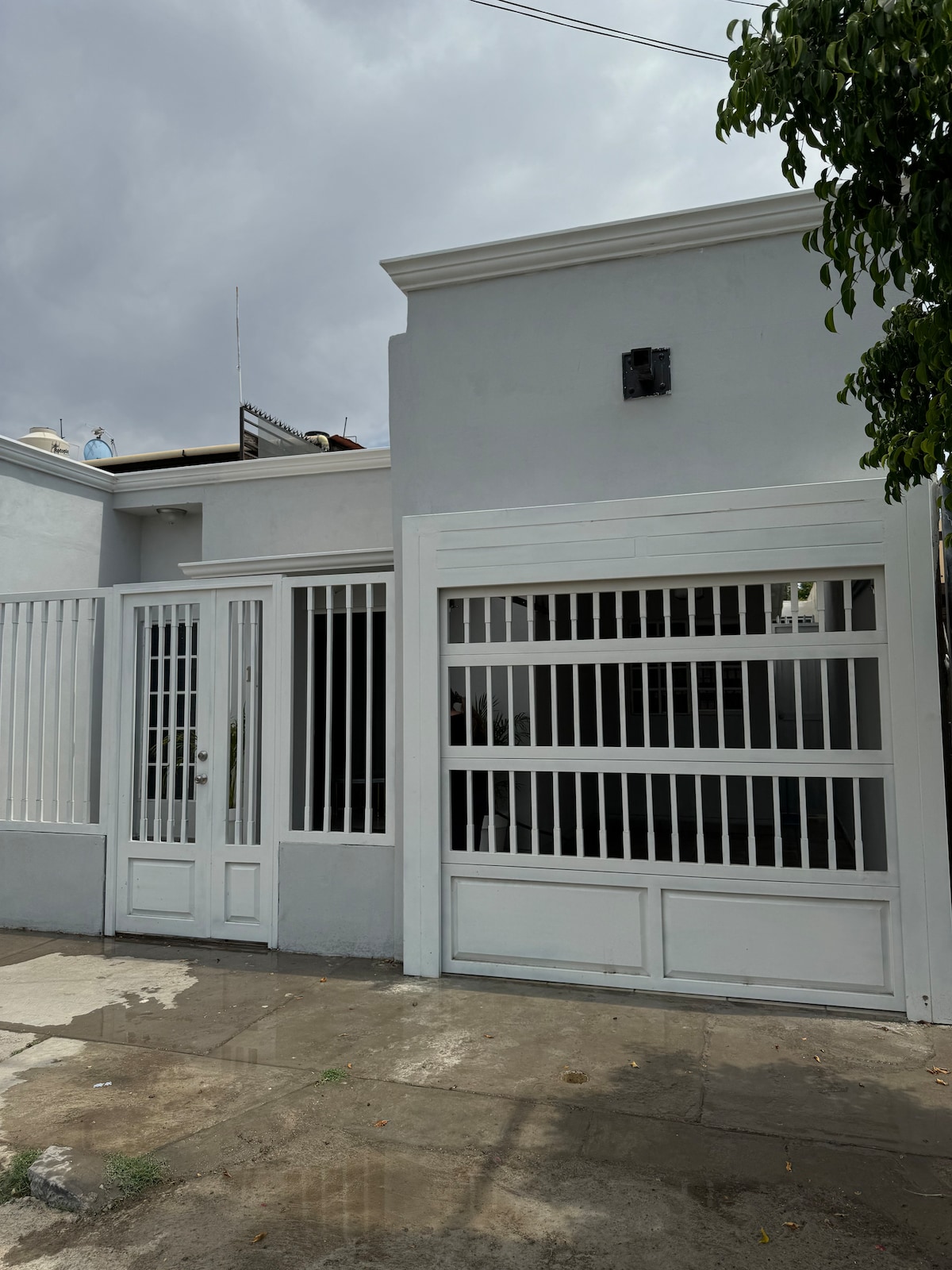 The exterior of the house features a modern design with light-colored walls and a gated entrance. The front is characterized by vertical white bars allowing visibility while providing privacy. A driveway is visible, leading to the garage located on the right side of the image.