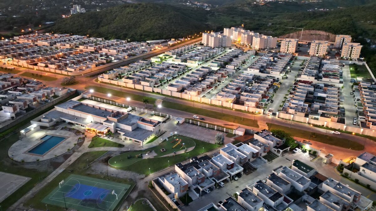 An aerial view captures a residential complex, showcasing neatly arranged houses and a swimming pool area. Nearby, green spaces and sports courts are visible, illuminated in the evening light. The surrounding hills create a natural backdrop, framing the community.
