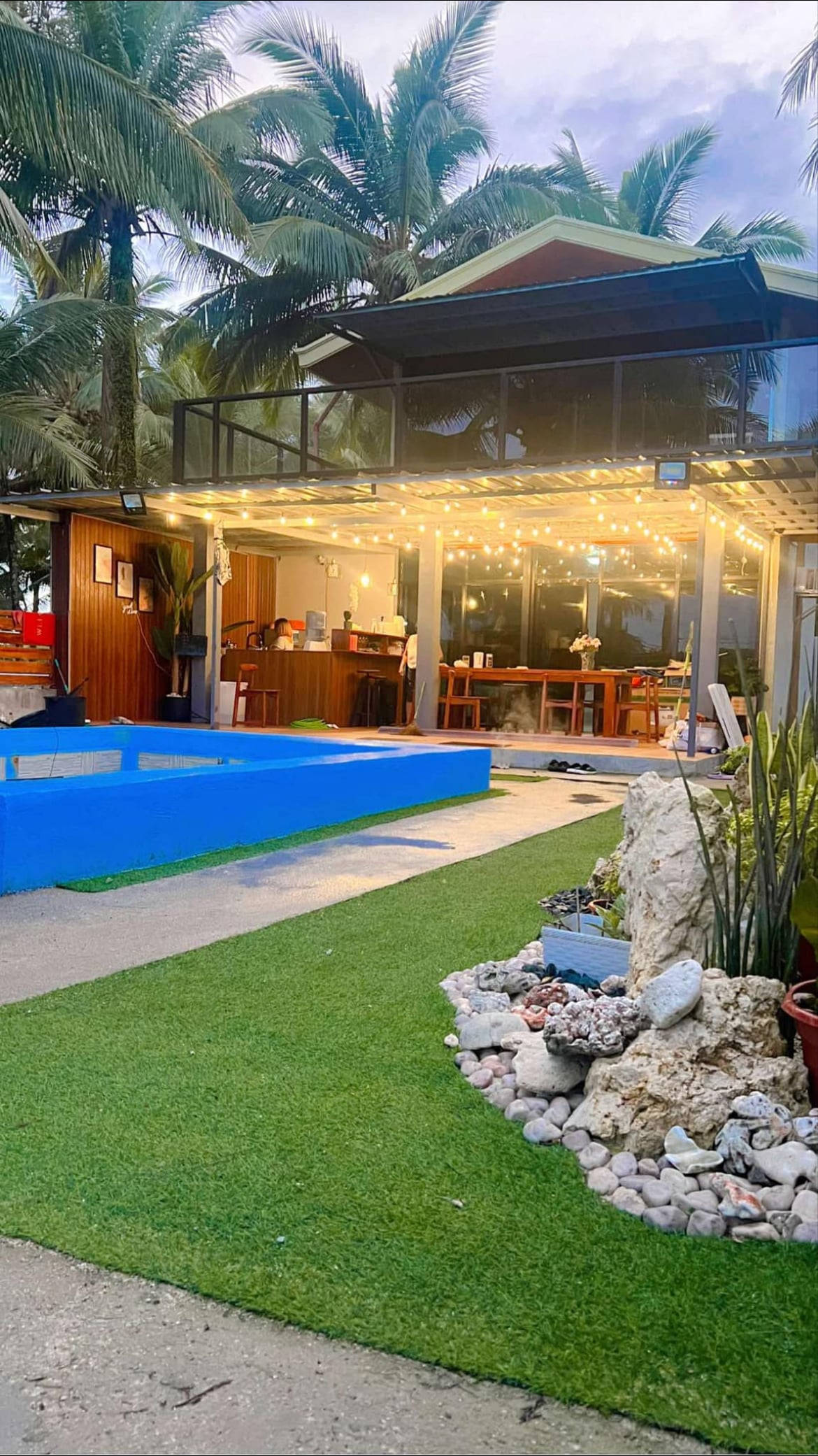 An outdoor area showcases a private saltwater pool beside a landscaped lawn. Soft lighting is draped above the patio, illuminating a cozy dining space with wooden furniture. Lush palm trees surround the area, while decorative stones and plants add texture to the environment.